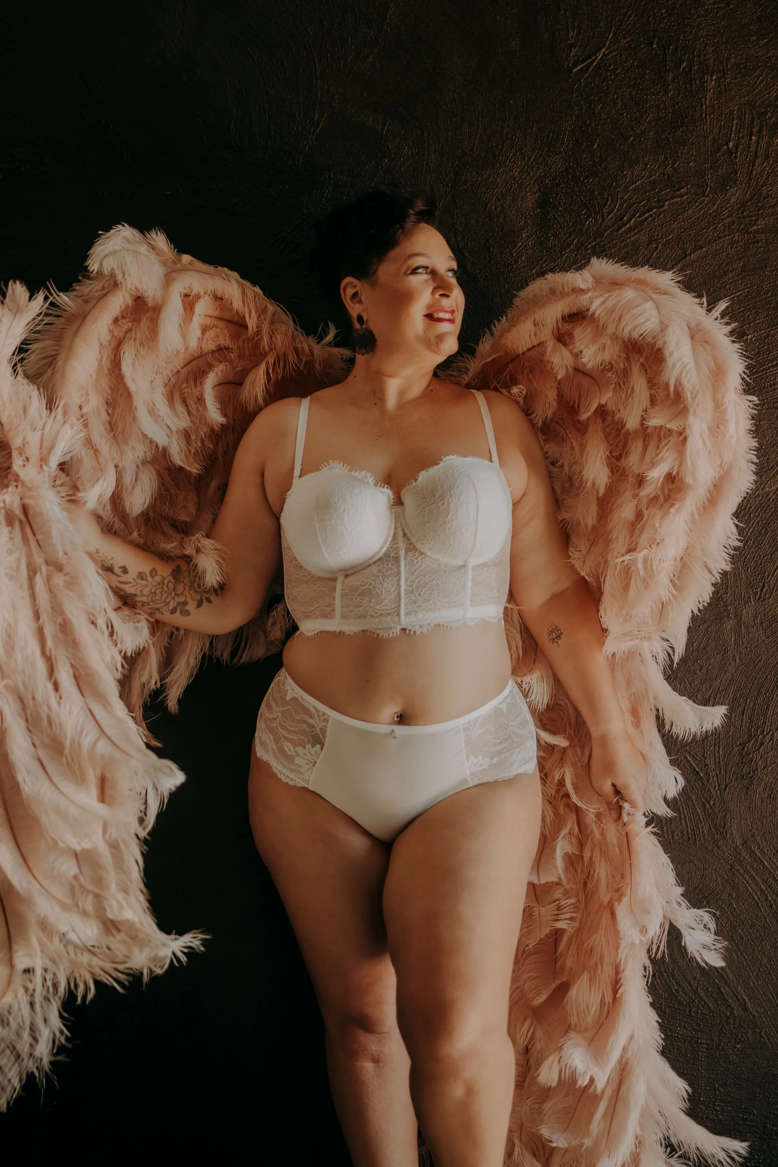 Woman lying on a black surface with pink feathered wings, wearing white lingerie, smiling and looking to the side.