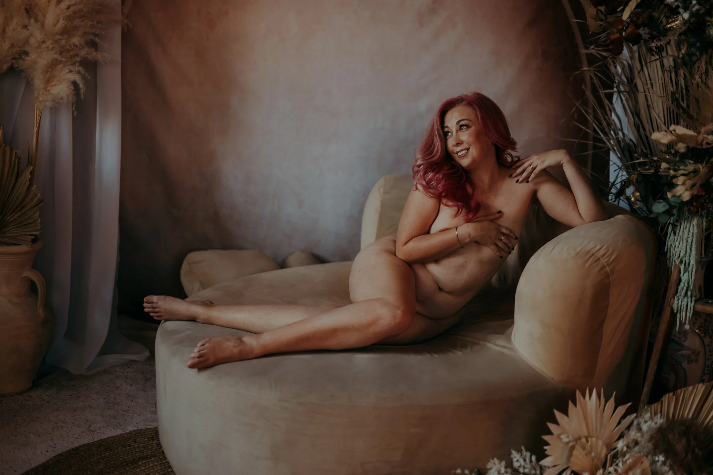 A woman with pink hair reclining nude on a beige velvet sofa, surrounded by dried floral arrangements in a softly lit room.