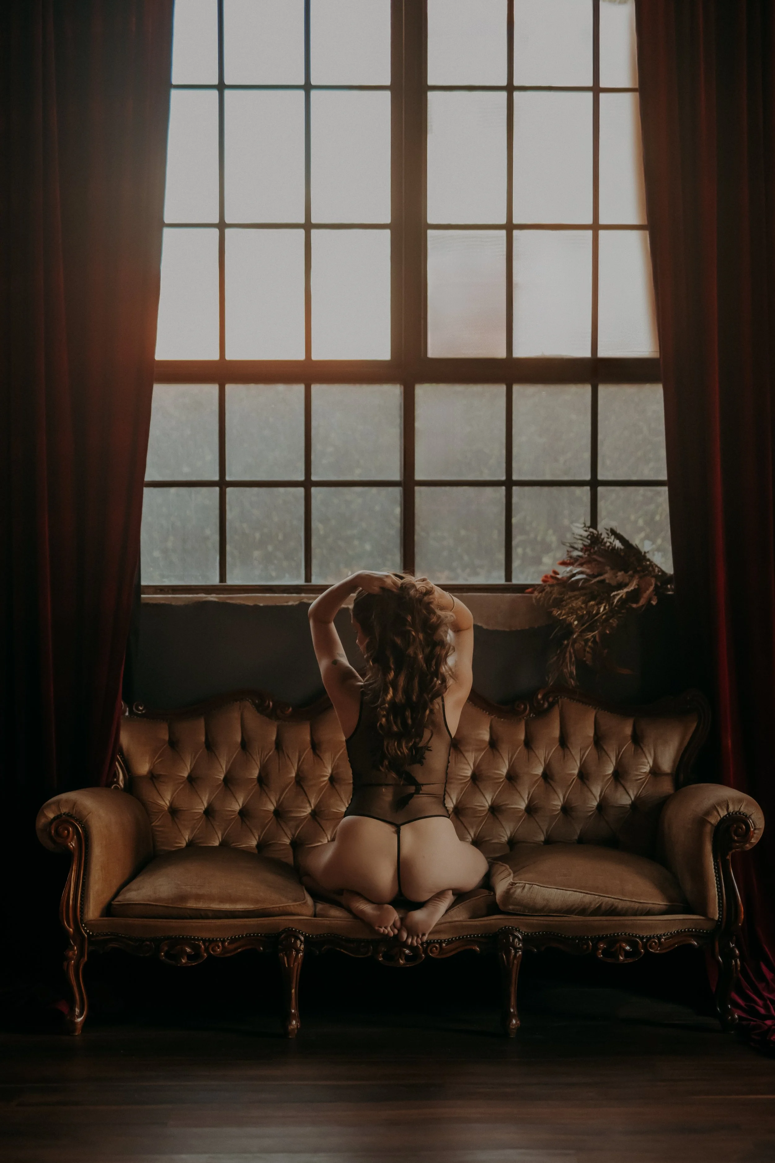 A woman in black lingerie kneels on a vintage-style sofa in front of large window with red curtains, with her back facing the camera and her hands in her hair.