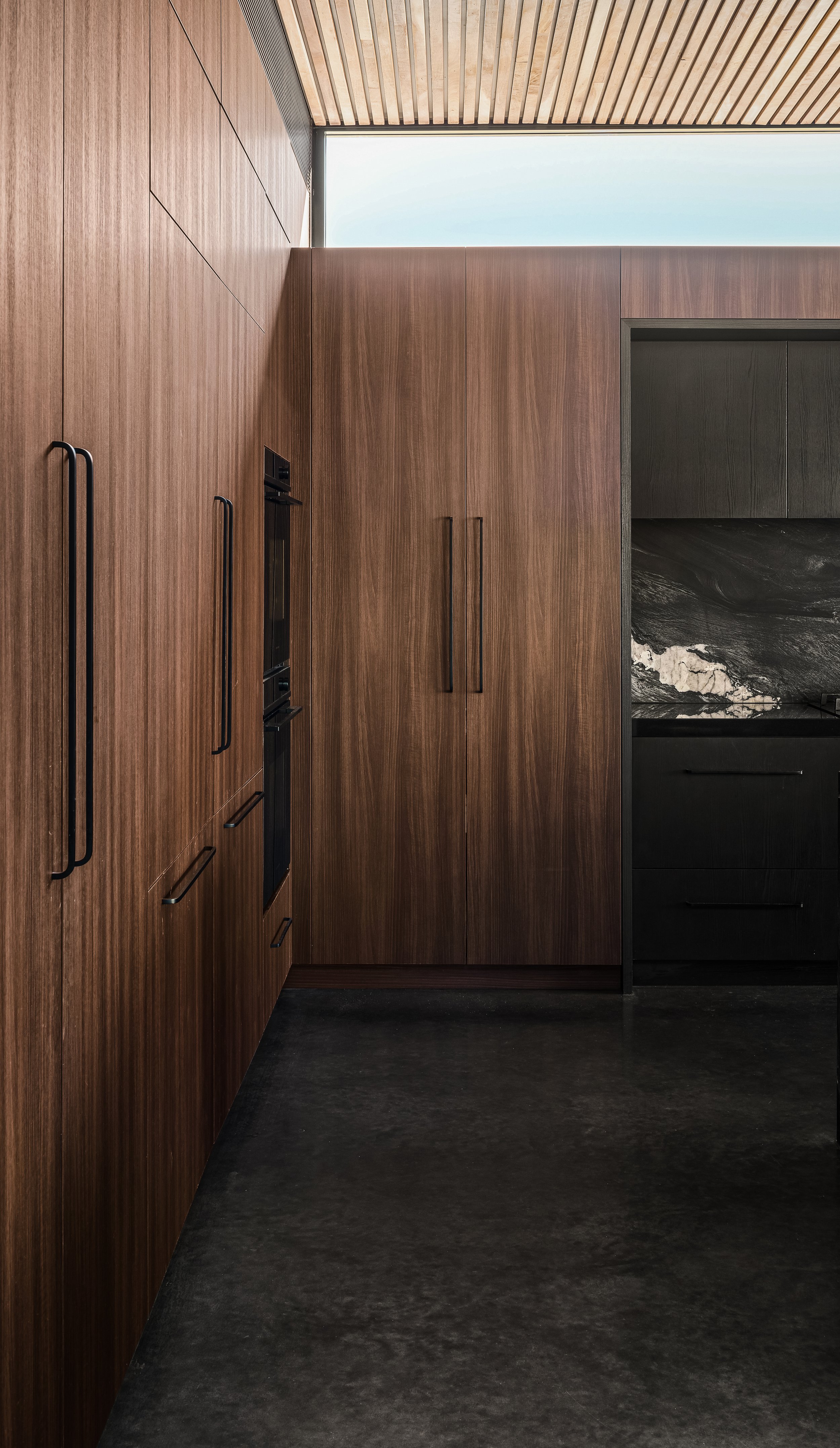 Modern kitchen with wooden cabinets and black appliances, dark flooring, and a large window with a wood ceiling.