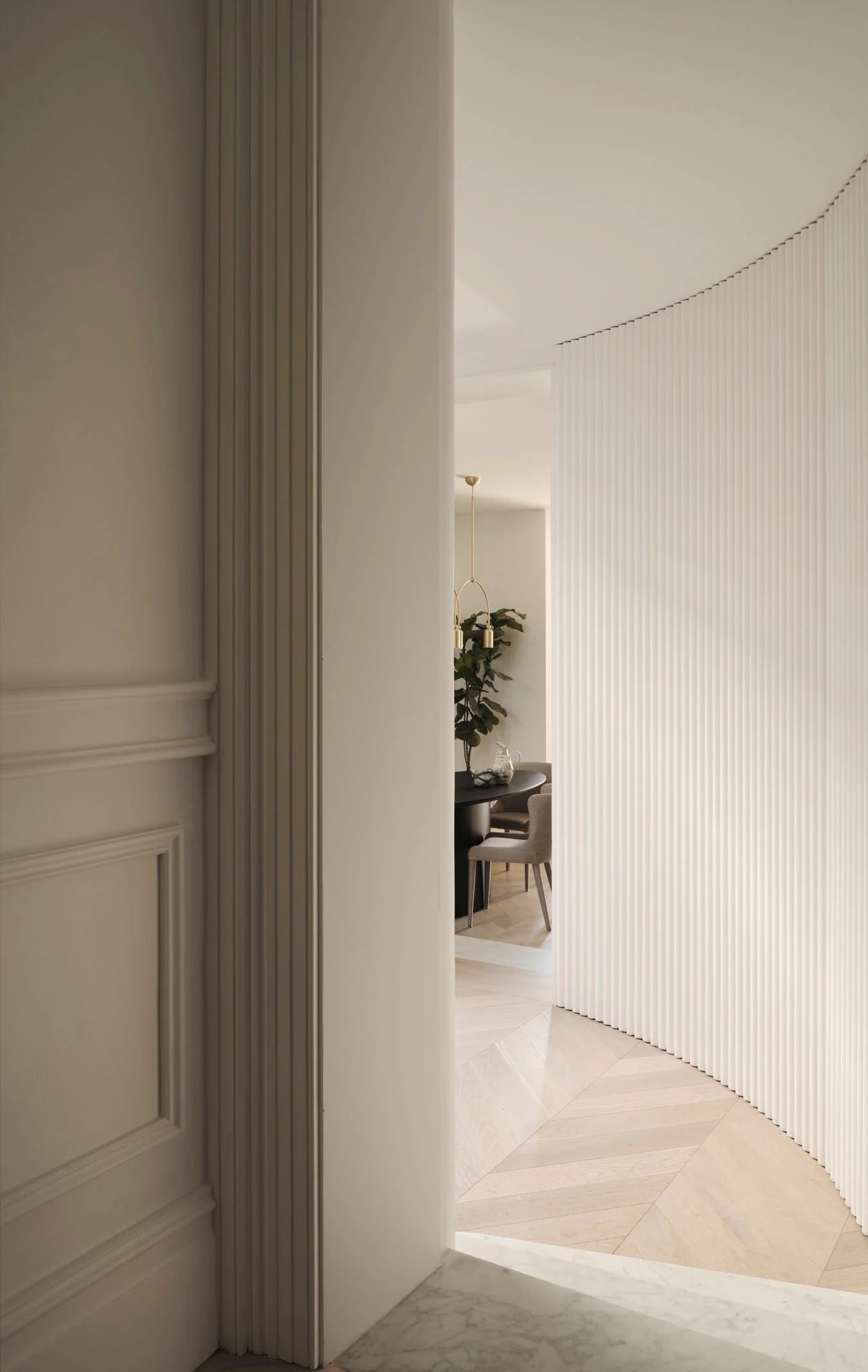An interior space with a partial view of a room through an open door. The room has a black table, chairs, a potted plant, and a hanging pendant light. The flooring is light-colored marble, and a partition or room divider with vertical grooves separat
