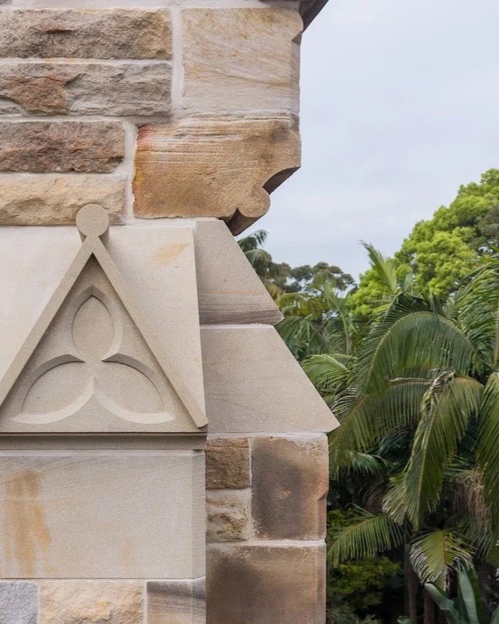 Close-up of a stone building corner with decorative triangular and curved stonework, and green trees in the background.