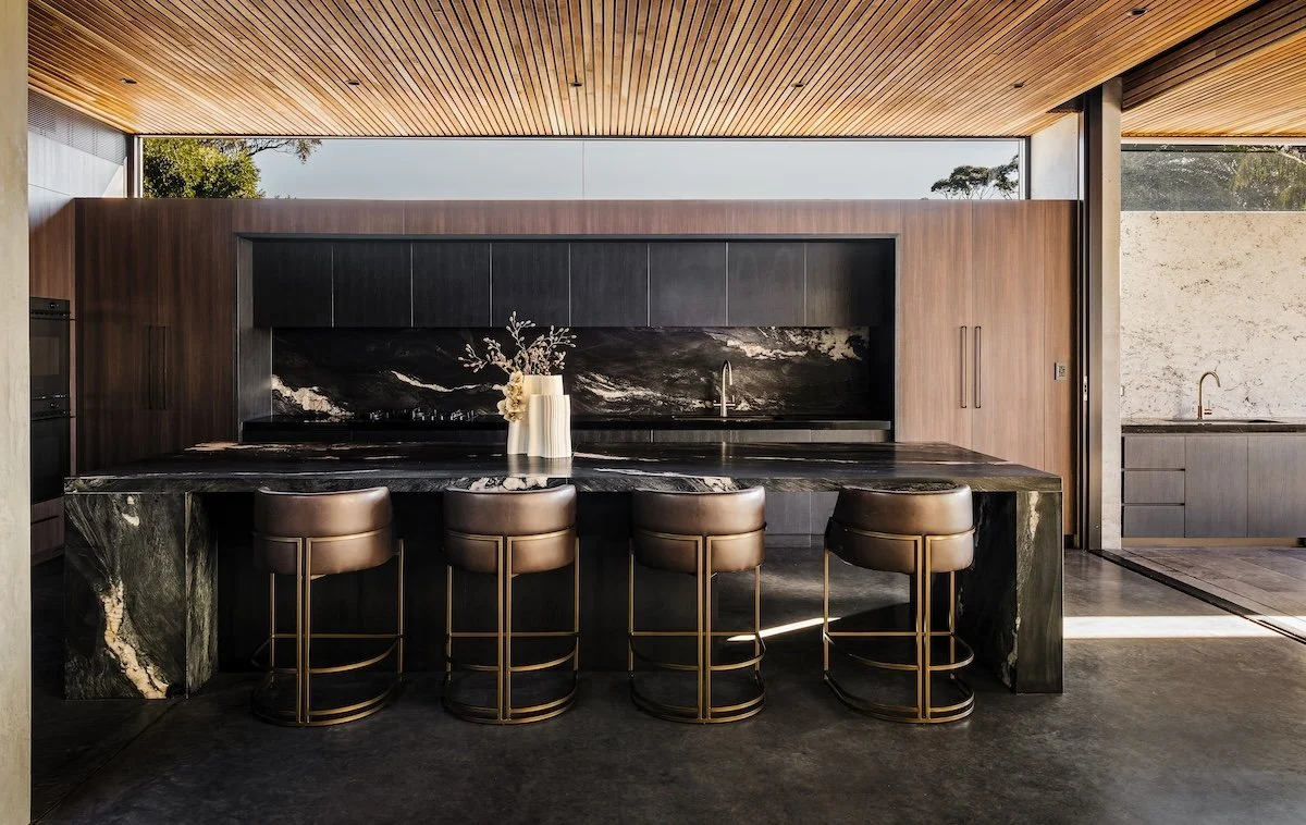 Modern kitchen featuring a black marble island with gold-accented bar stools, black upper cabinets, and a wood ceiling. There is a vase with flowers on the island and a large window providing natural light.