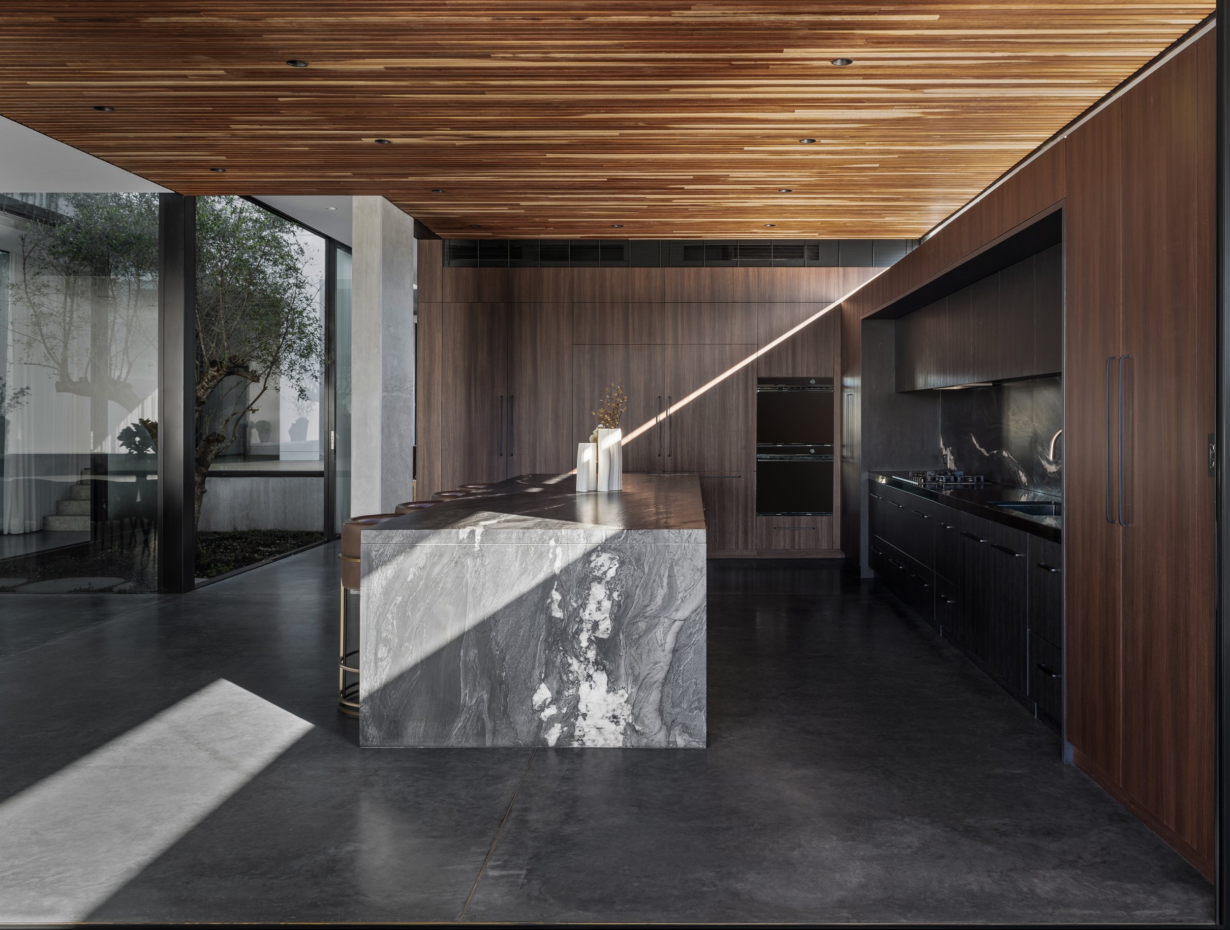 Modern kitchen with dark wood cabinets, marble island, and large glass window showing trees outside.