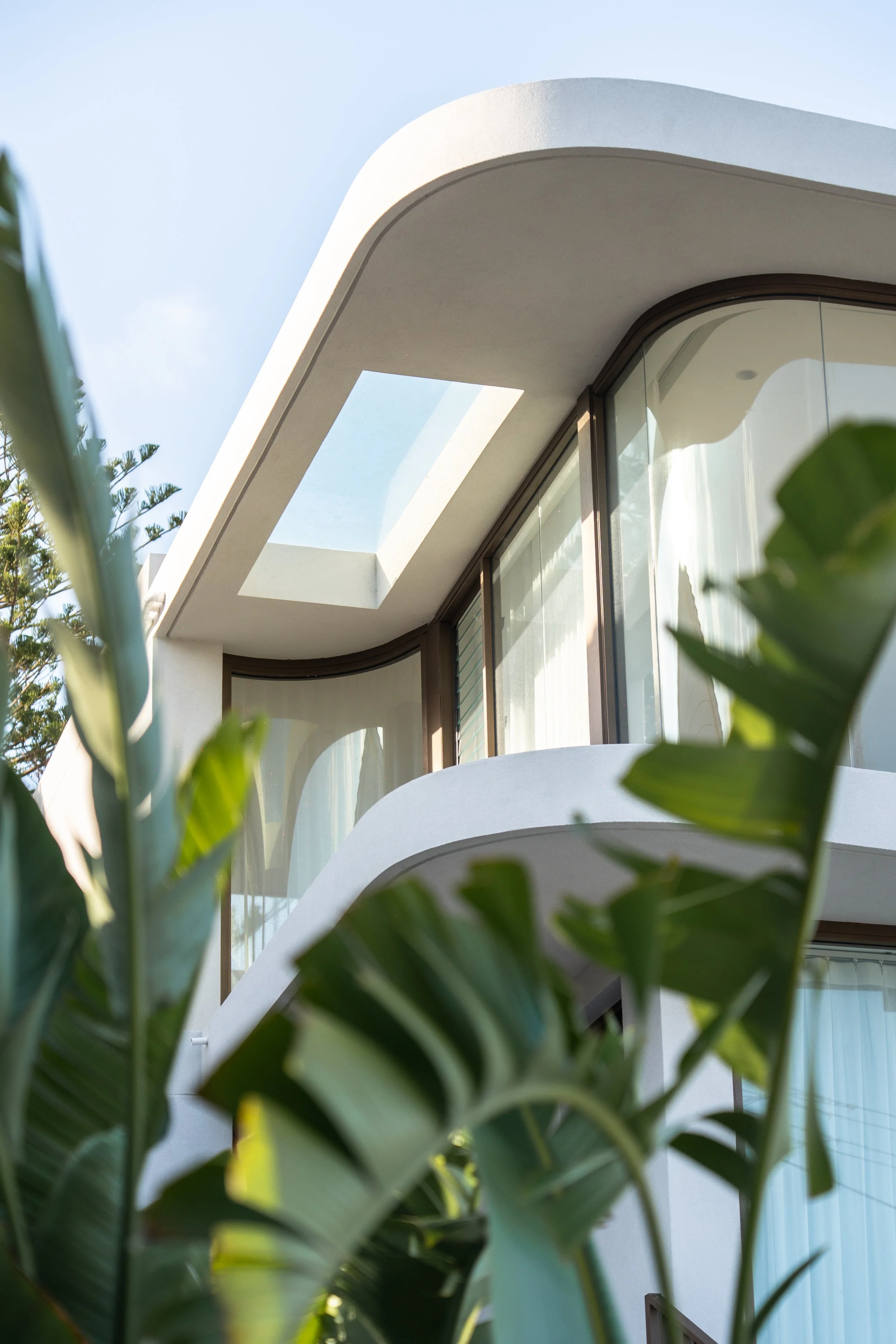 Modern white building with curved balconies and large windows, surrounded by green plants.