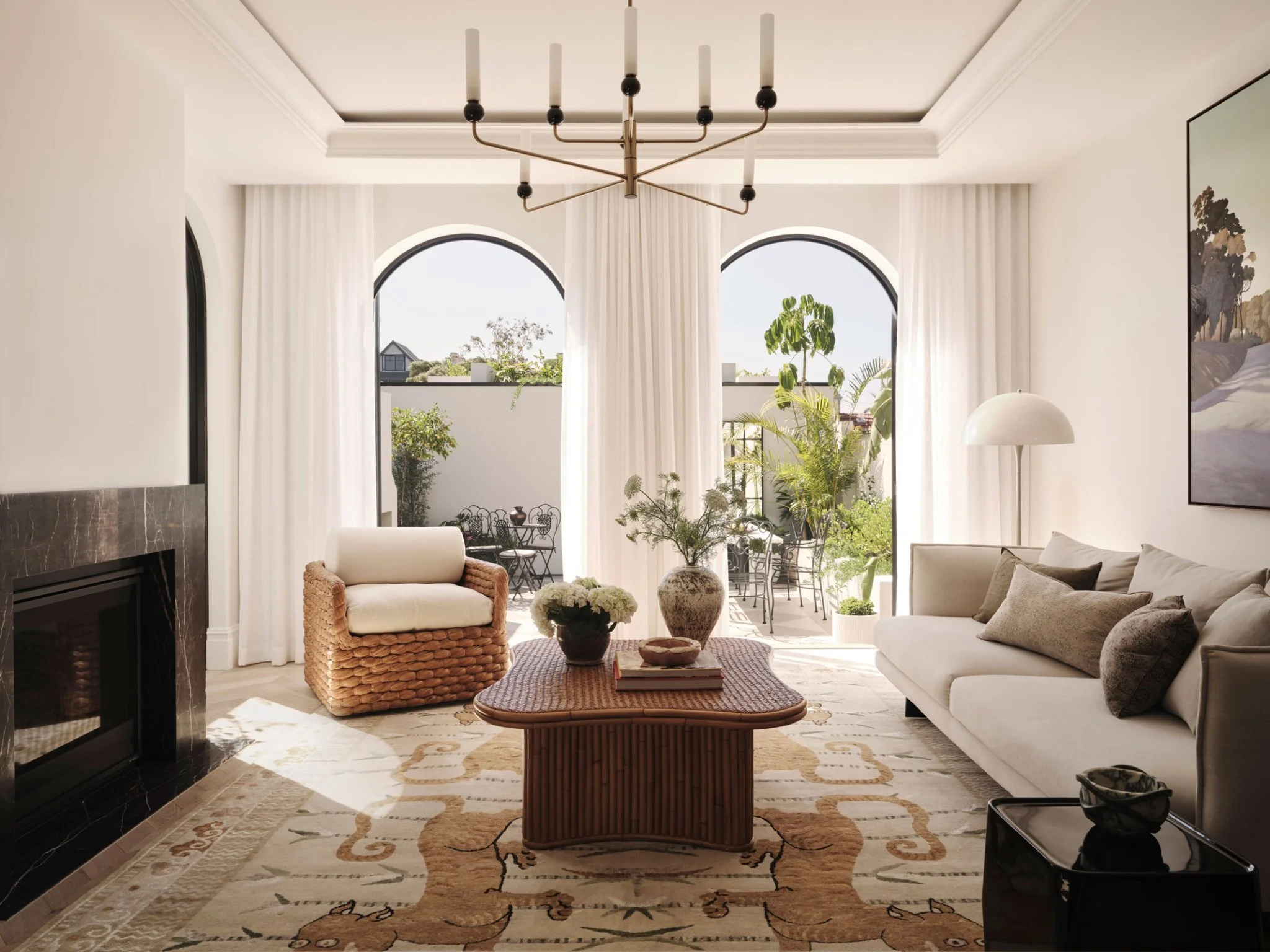 A bright living room with large arched windows, white curtains, and a view of greenery outside. The room features a white sofa, a wicker armchair, a coffee table with books and decorative items, and various plants. There is a modern chandelier hangin