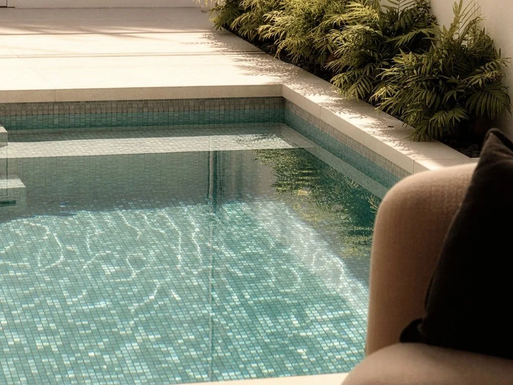 Close-up view of a small swimming pool with turquoise tiles, surrounded by a white concrete deck and lush green plants on one side, with sunlight casting shadows.