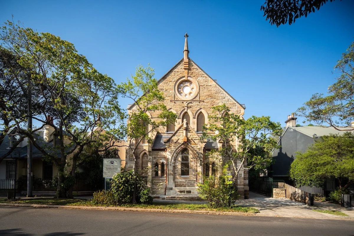 Front view of a historic stone church with a large circular window and pointed arches, surrounded by trees and residential houses under a clear blue sky.