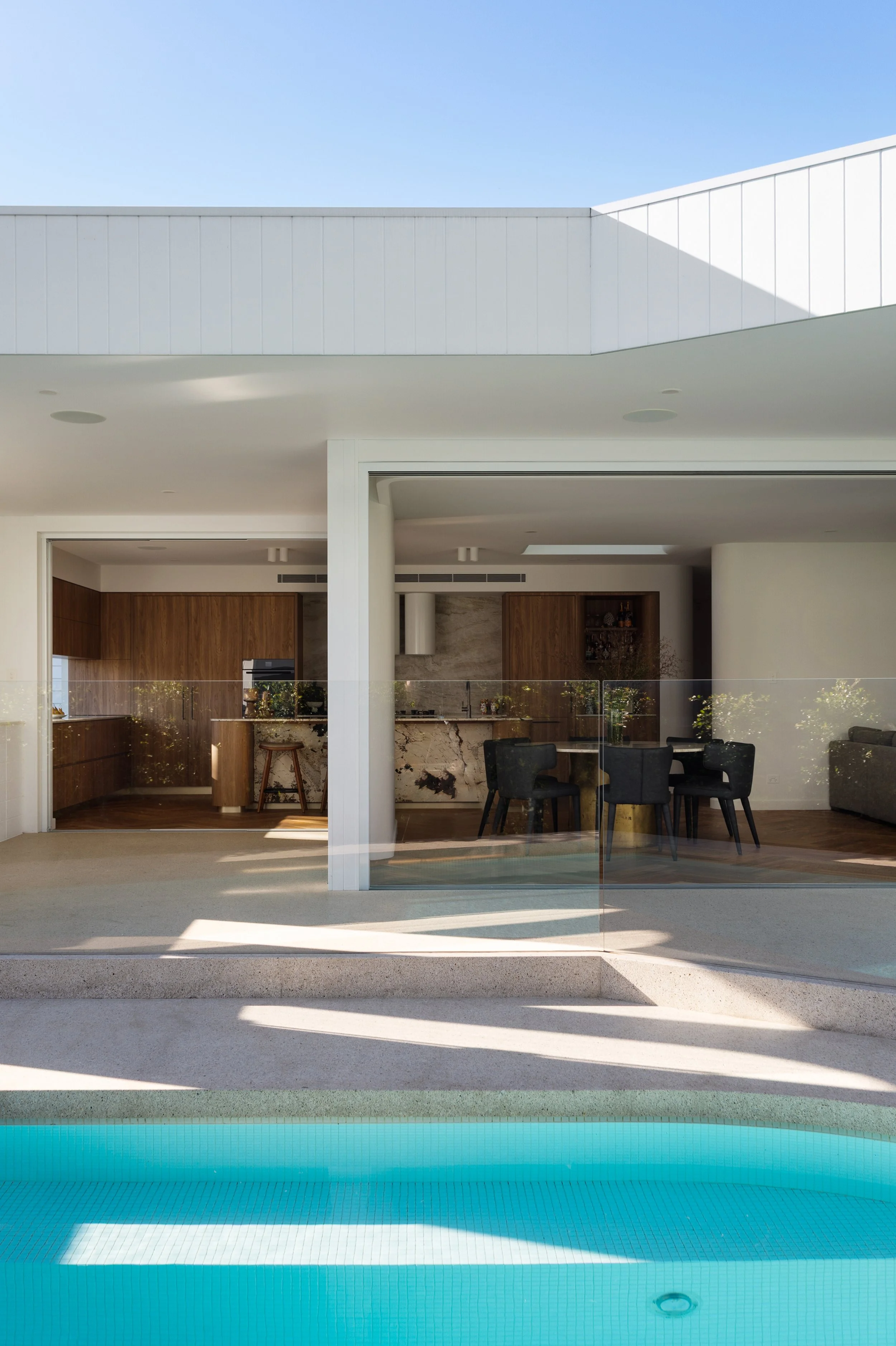 Modern house with large glass sliding doors opening to a backyard with a swimming pool. The house features white walls, wood accents, and a minimalist design. Clear blue sky is visible above.