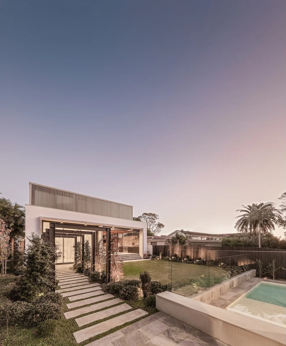 Modern backyard with a landscaped garden, a pathway leading to a house with a large open window, a swimming pool with glass fencing, and palm trees against a twilight sky.