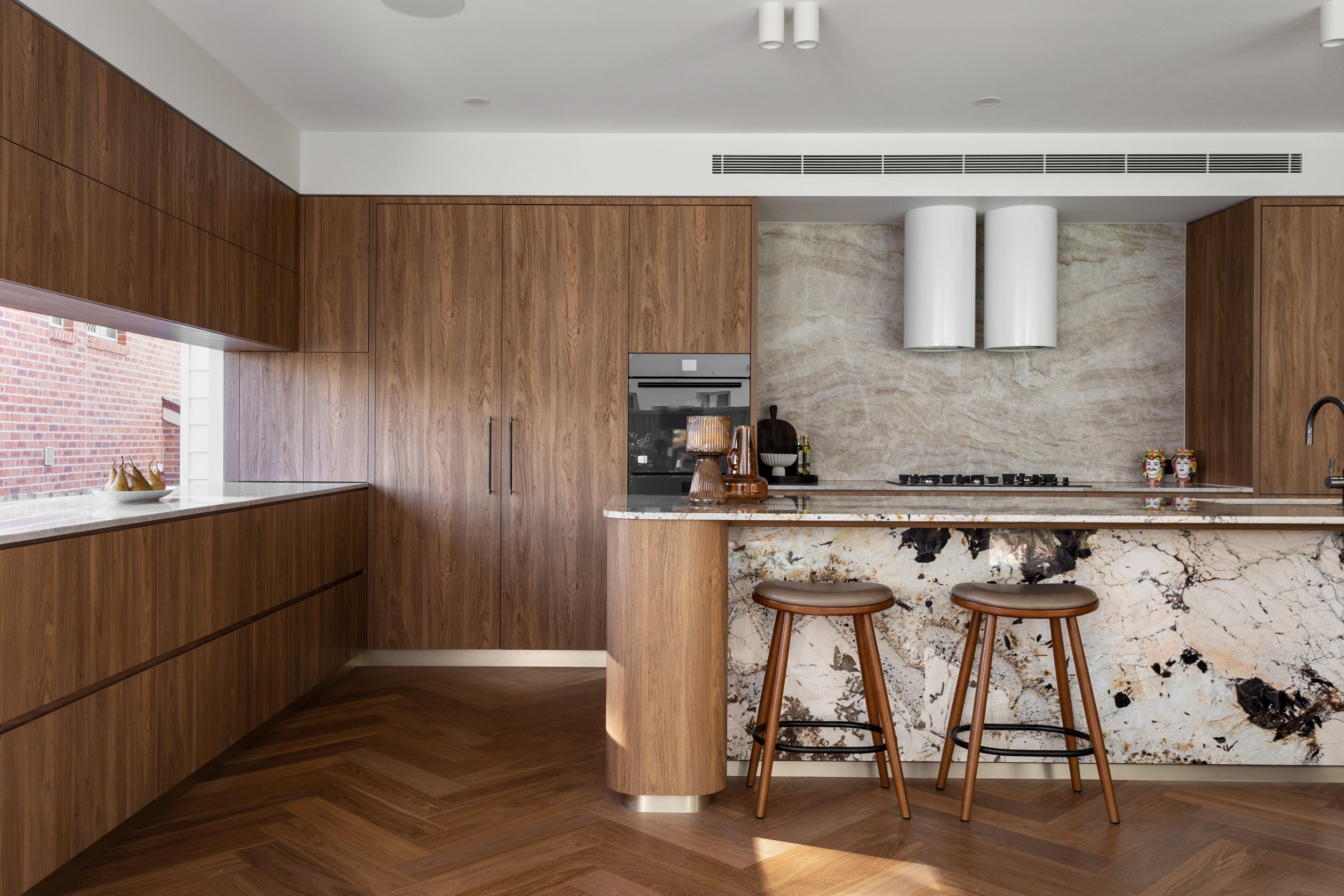 Modern kitchen with wooden cabinets, a marble countertop, bar stools, and decorative items, including a ceramic pair and art pieces, against a marble backsplash with a gas stove.