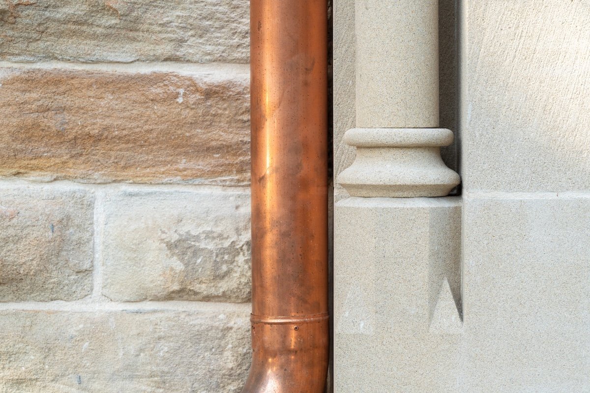 Close-up of a copper rain gutter pipe against a brick wall and a beige stone building.