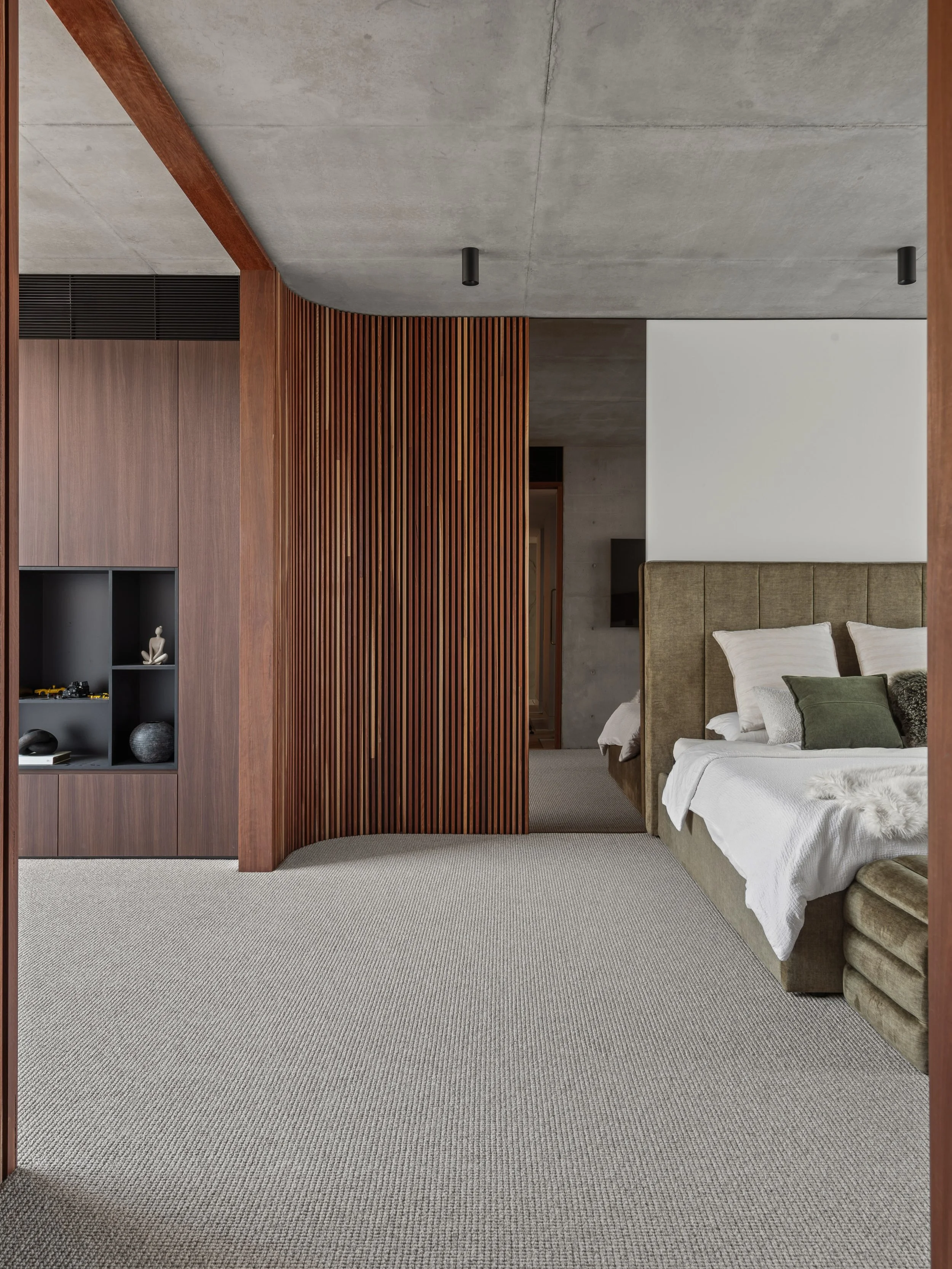 A bedroom with a beige upholstered bed, white and green pillows, a grey carpet, a wooden slat room divider, and a concrete ceiling with black ceiling lights.