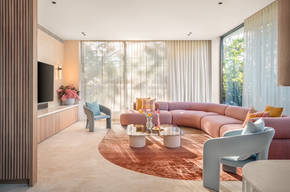 Bright living room with large windows, pink sectional sofa, light blue armchairs, round glass coffee table, and beige curtains.