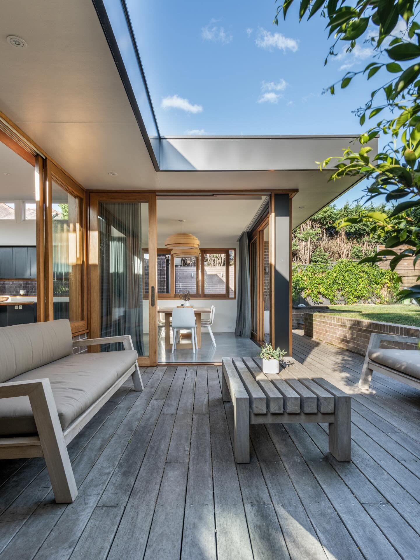 View of a modern outdoor patio area with wooden flooring, white outdoor sofas, a wooden coffee table, and sliding glass doors leading into a dining room with a white table, chairs, and a pendant light. The patio is open to a garden with green trees a