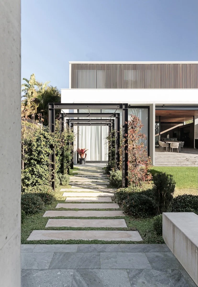Modern house with a landscaped pathway leading to a patio with outdoor dining area, greenery, and a tall palm tree, under a clear blue sky.