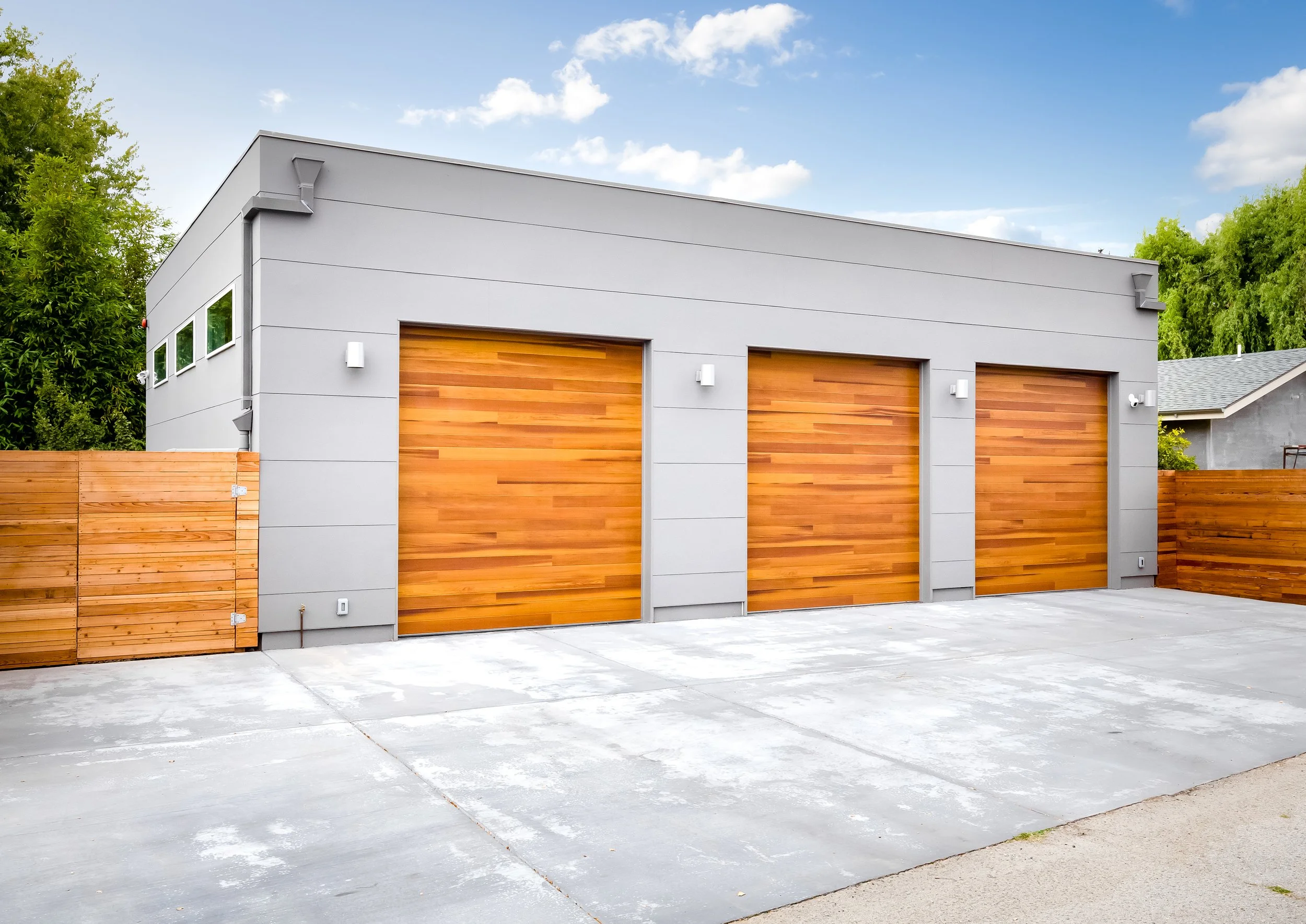 A modern detached garage with three wooden garage doors, gray exterior walls, and a concrete driveway, with a blue sky and some trees in the background. New garage build in Santa Cruz, CA. 