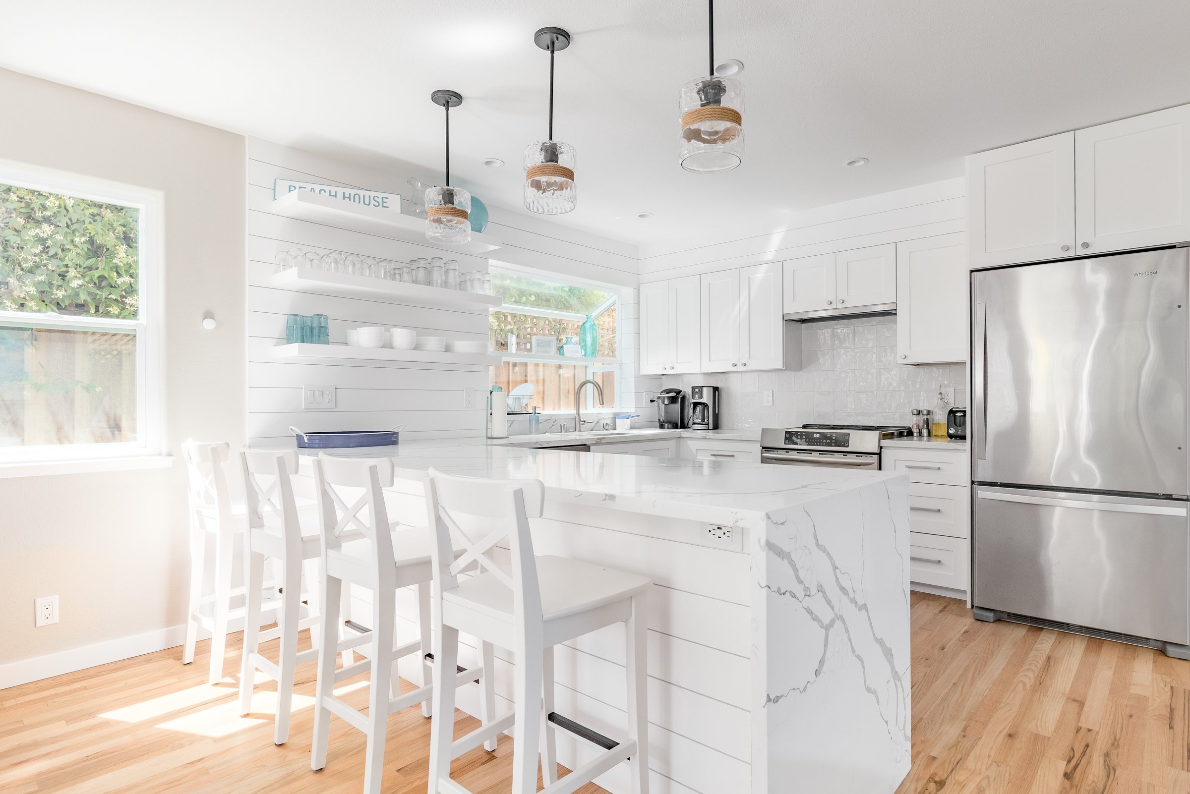 Bright modern kitchen with white cabinets, marble island, and light wood flooring, featuring pendant lights and large windows.