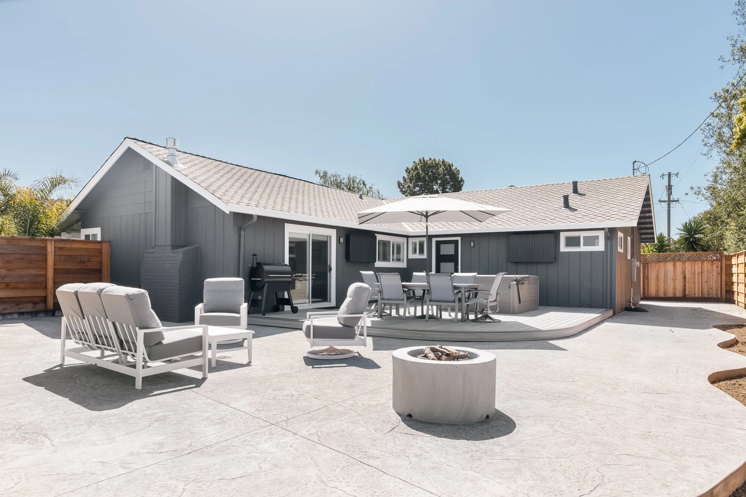 Backyard patio with outdoor furniture, umbrella, fire pit, and a gray house with sliding glass door, wooden fence, and utility poles under a clear sky. Concrete patio and outdoor hardscape in Santa Cruz, CA.