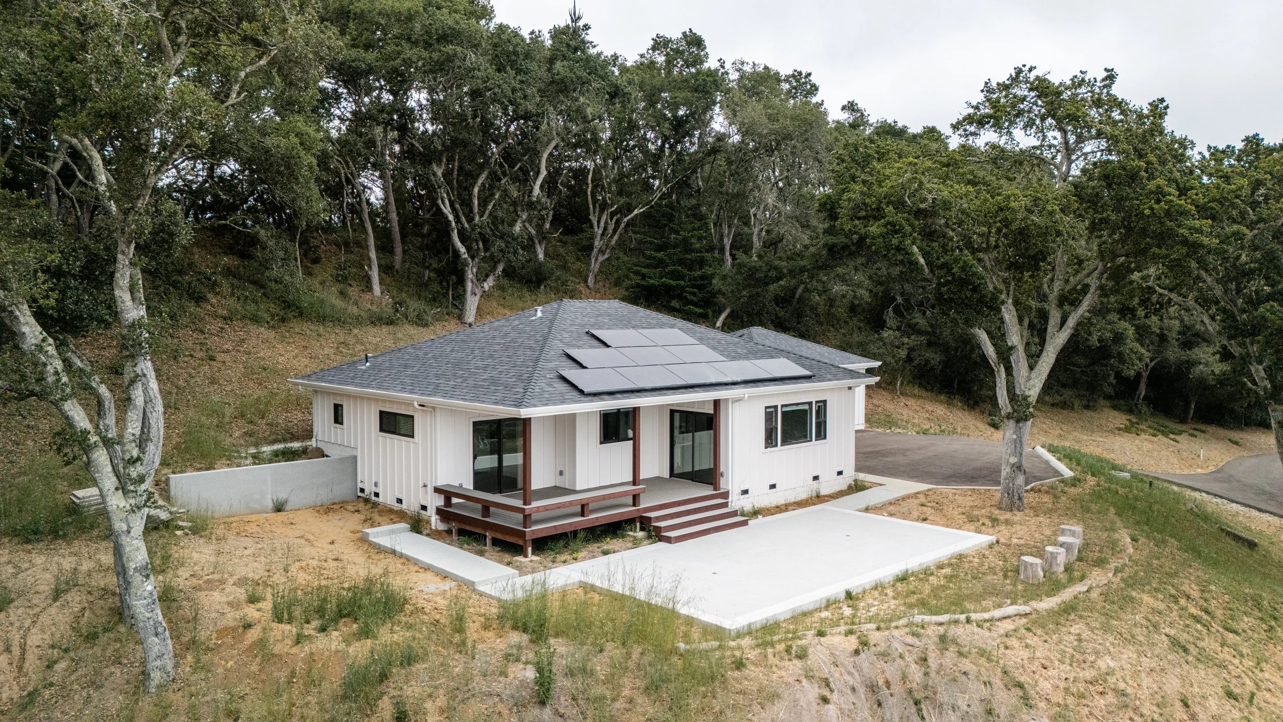 A modern single-story house with a gray roof and solar panels, white exterior walls, a small porch with wooden railing, large windows, and a concrete patio, surrounded by trees and natural landscape. Accessory Dwelling Unit (ADU) in Aptos, CA.