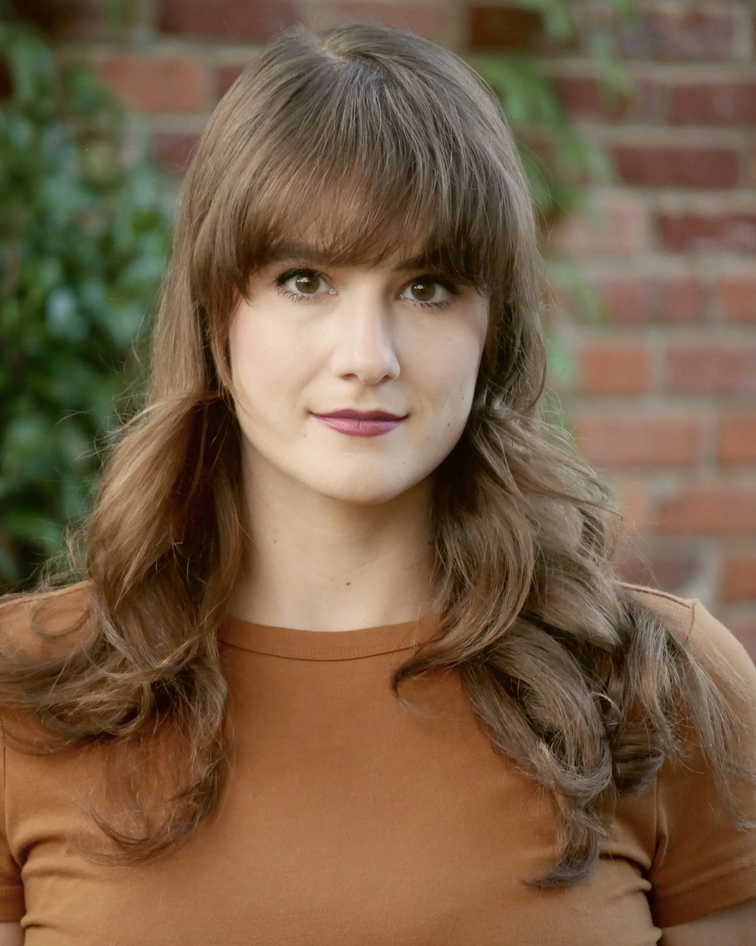 A young woman with wavy brown hair and bangs, wearing a brown top, standing outdoors in front of a brick wall and greenery.