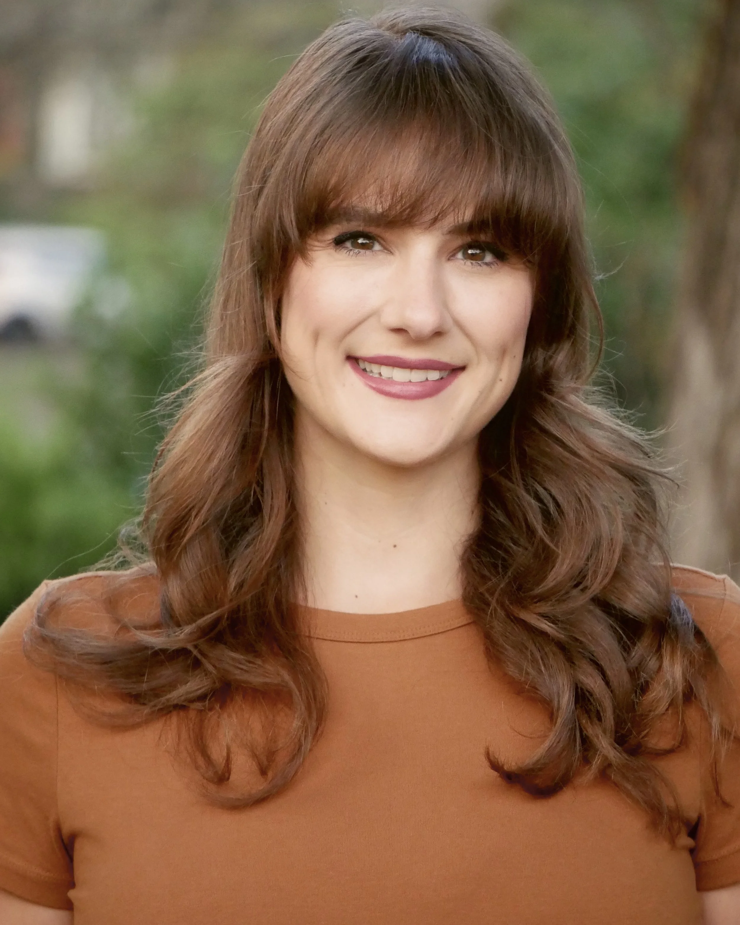 A woman with brown hair, wearing a rust-colored shirt, smiling outdoors with blurred greenery and trees in the background.