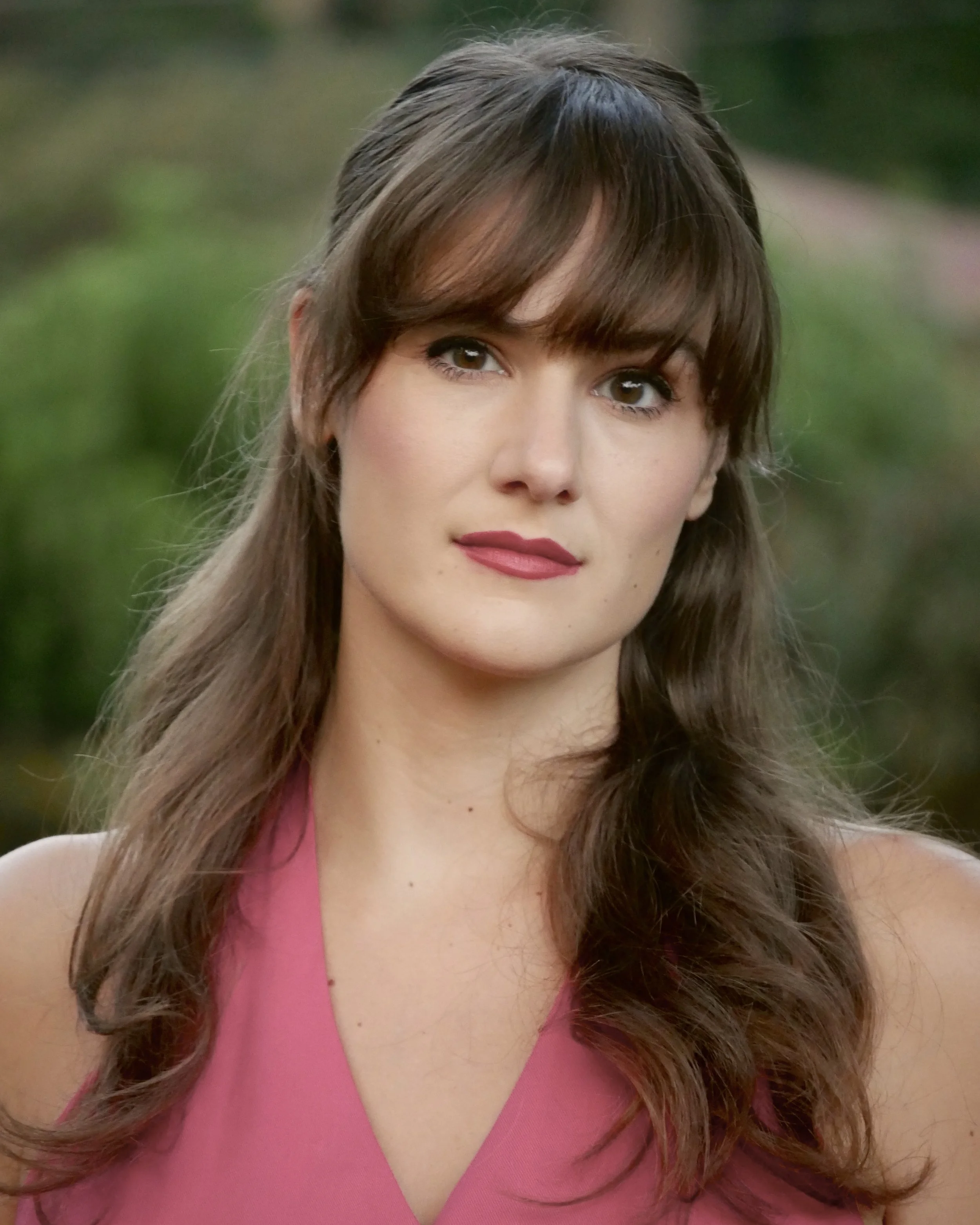 Close-up of a woman with long brown hair and bangs, wearing a pink sleeveless top, standing outdoors with blurred greenery in the background.