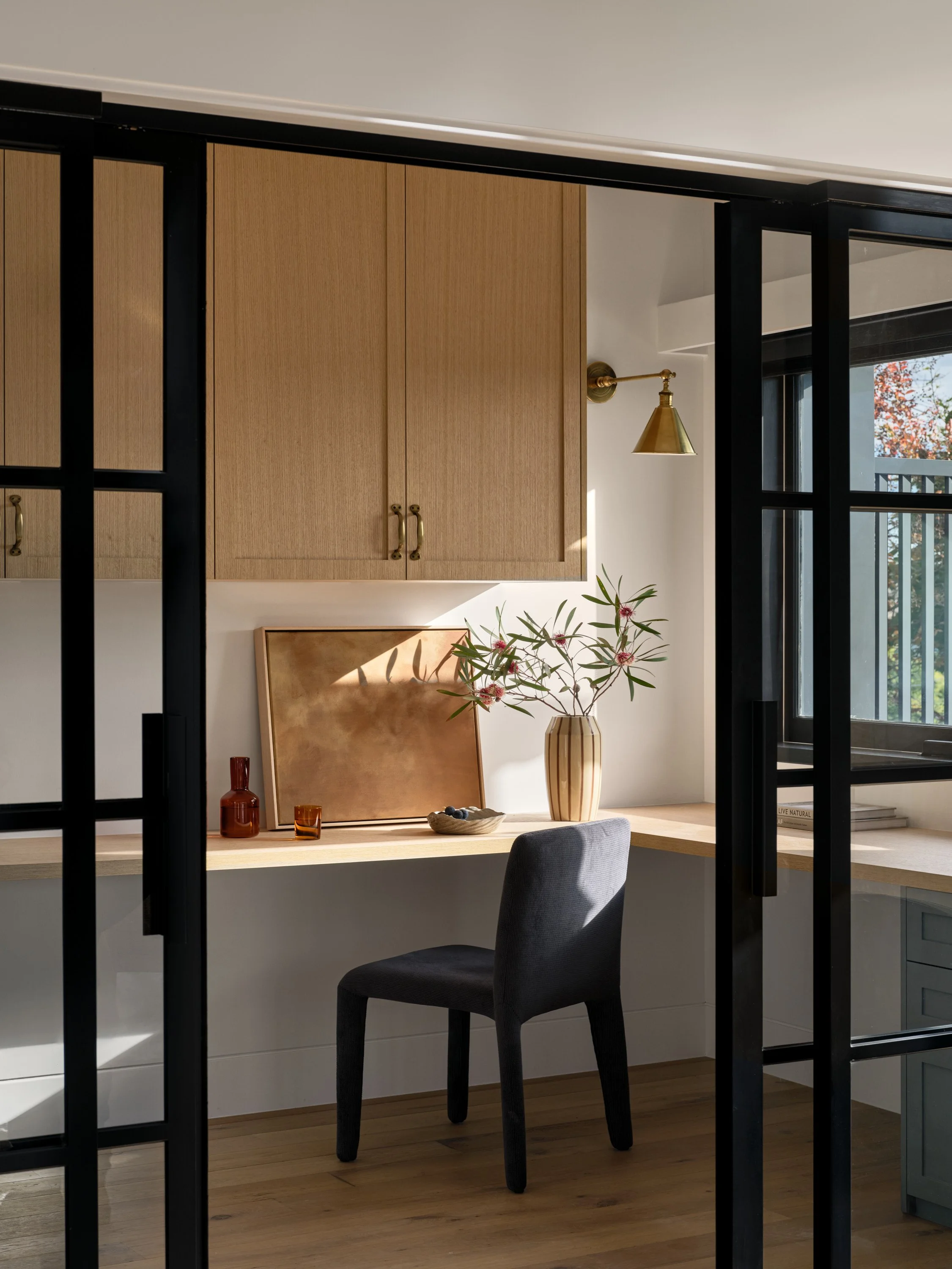 A modern home office with a wooden desk, a gray upholstered chair, large window with natural light, and decorative items including vases and framed artwork.