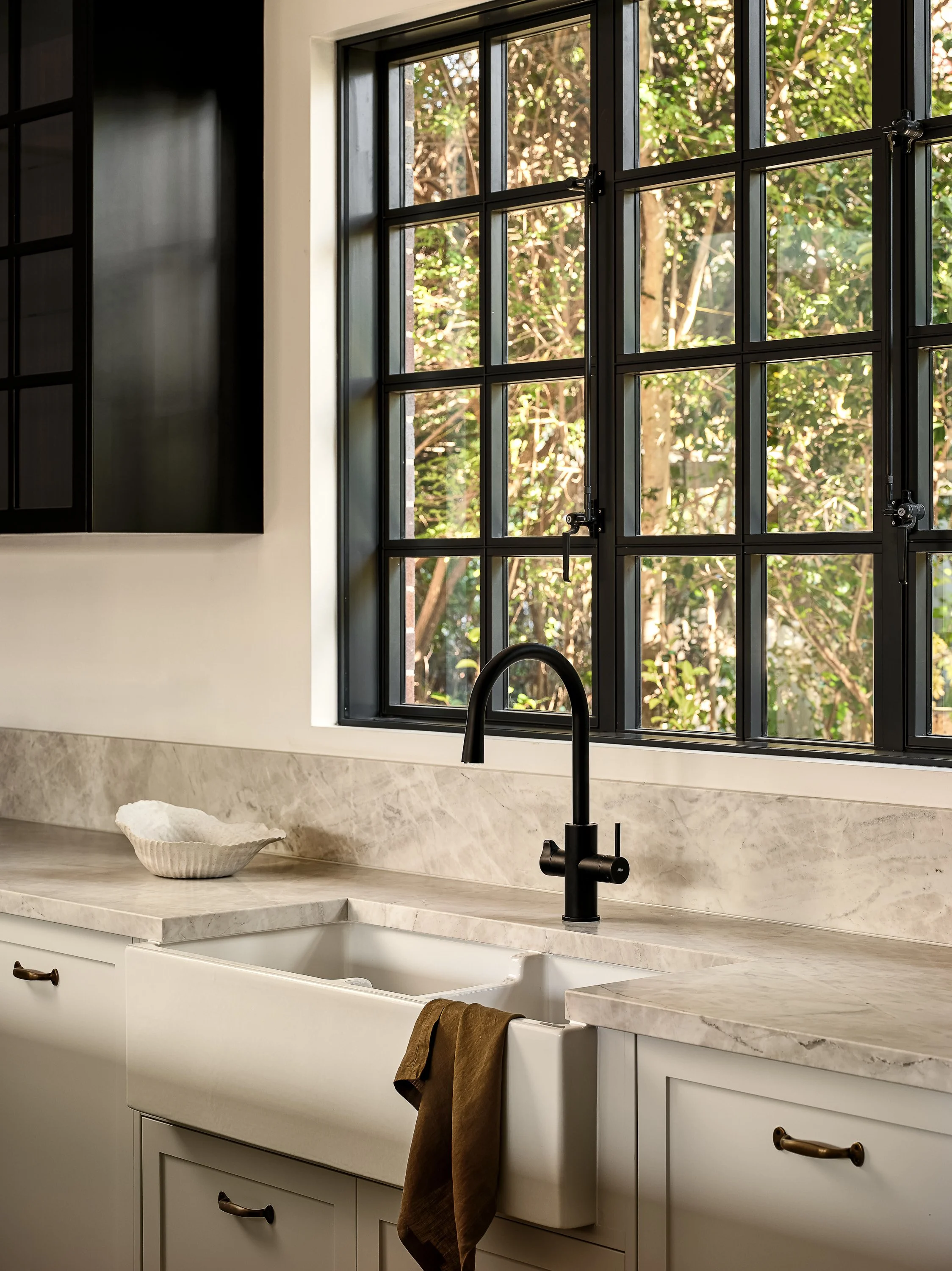 Kitchen sink with black faucet, white cabinetry, marble countertop, window with black frame showing trees outside, brown towel hanging from sink.