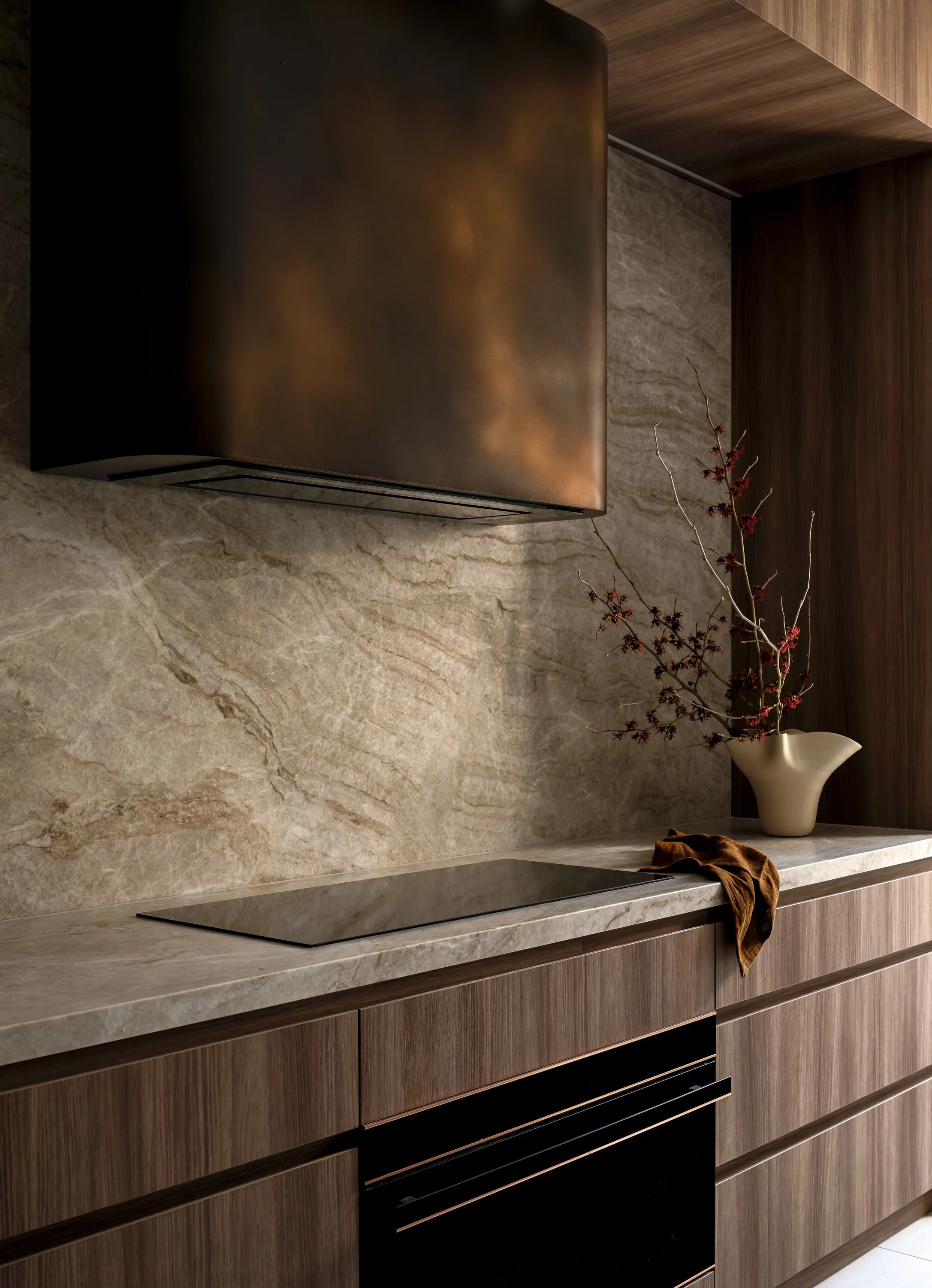 Modern kitchen with wooden cabinets, beige marble countertop, built-in black cooktop, decorative vase with dried branches, and a brown cloth on the counter.