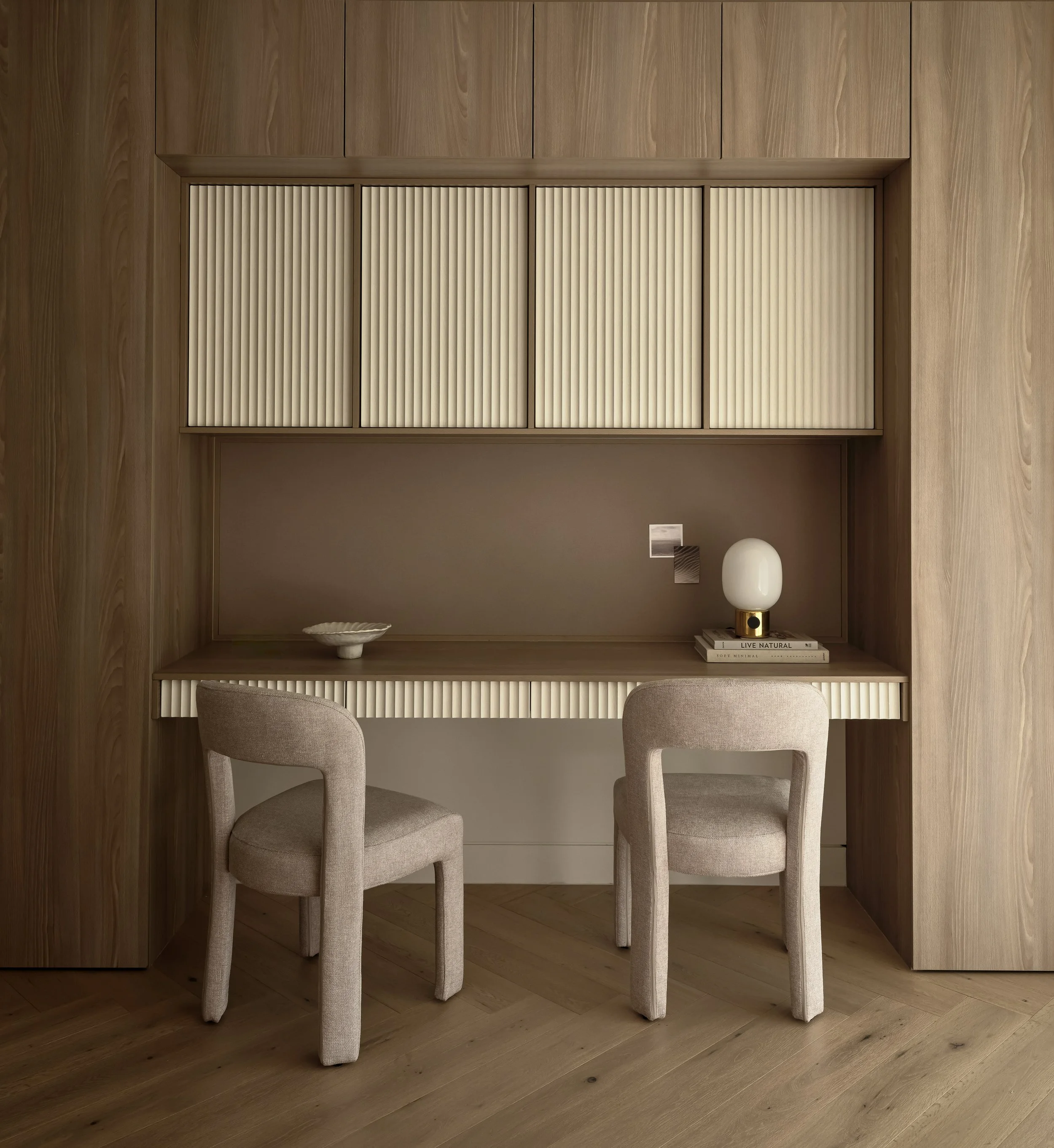 Interior space with wood-paneled walls and built-in shelves. Two beige upholstered chairs are positioned in front of a desk or table with a white ceramic bowl, a stack of books, and a decorative white vase. The room has a minimalist, modern design.