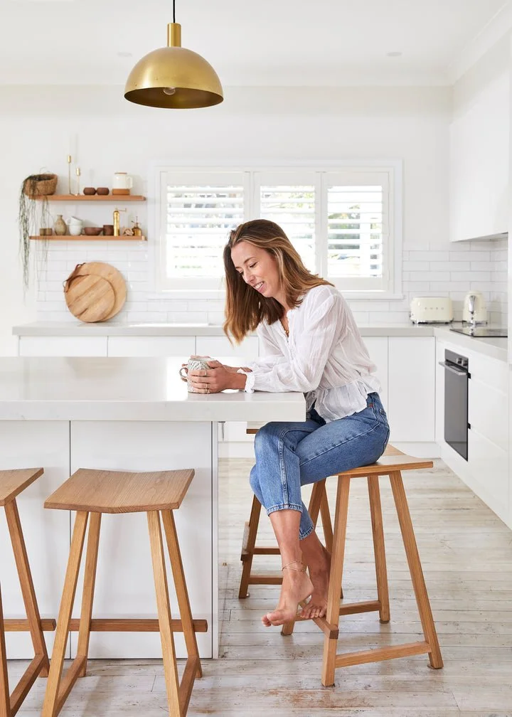 A woman sitting on a wooden bar stool in a bright, white kitchen, smiling while holding a mug. The kitchen features white cabinets, a white countertop, wooden open shelves with decor, and a large window with white shutters. There is a gold pendant li