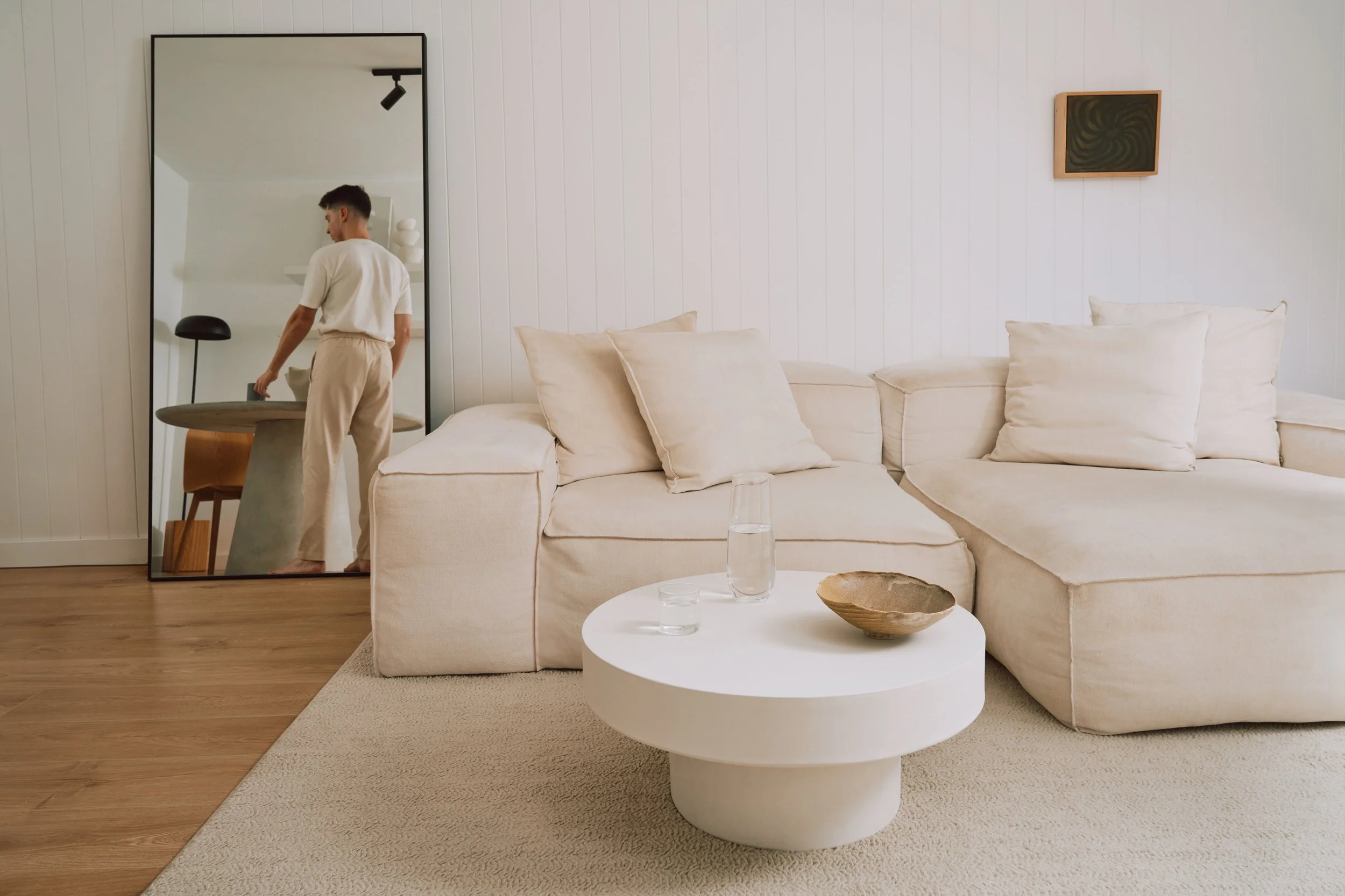 A living room with a beige sectional sofa, a white round coffee table with a bowl and water glass, a large mirror reflecting a person, and minimalist artwork on white paneled walls.
