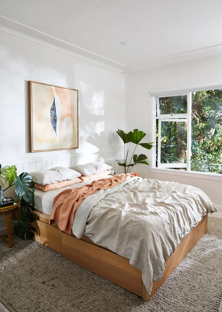 A bedroom with a bed, linen, and decorative plants near a window with a view of trees outside.