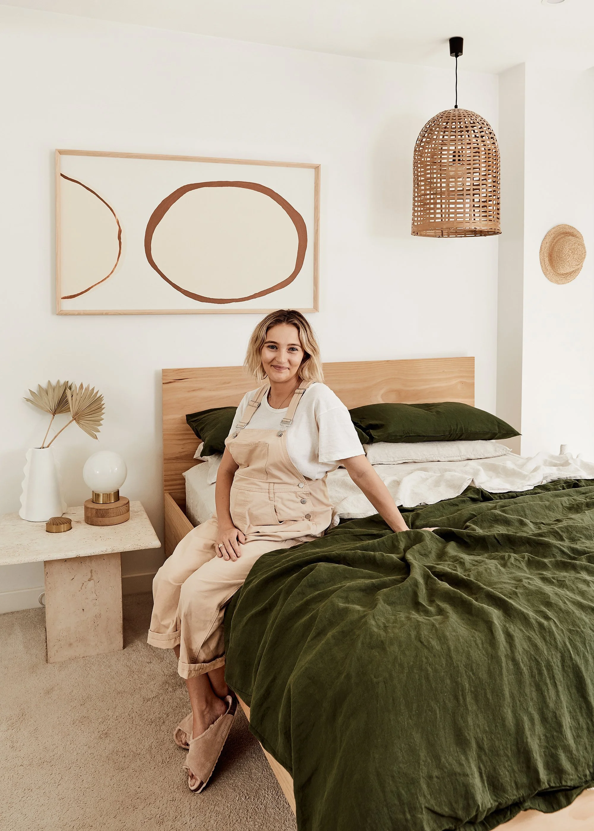 A woman with blonde hair sitting on the edge of a bed with dark green bedding in a modern, minimalistic bedroom. Behind her, there is a wooden headboard, a nightstand with a white vase and a small table lamp, and a large abstract artwork with brown o