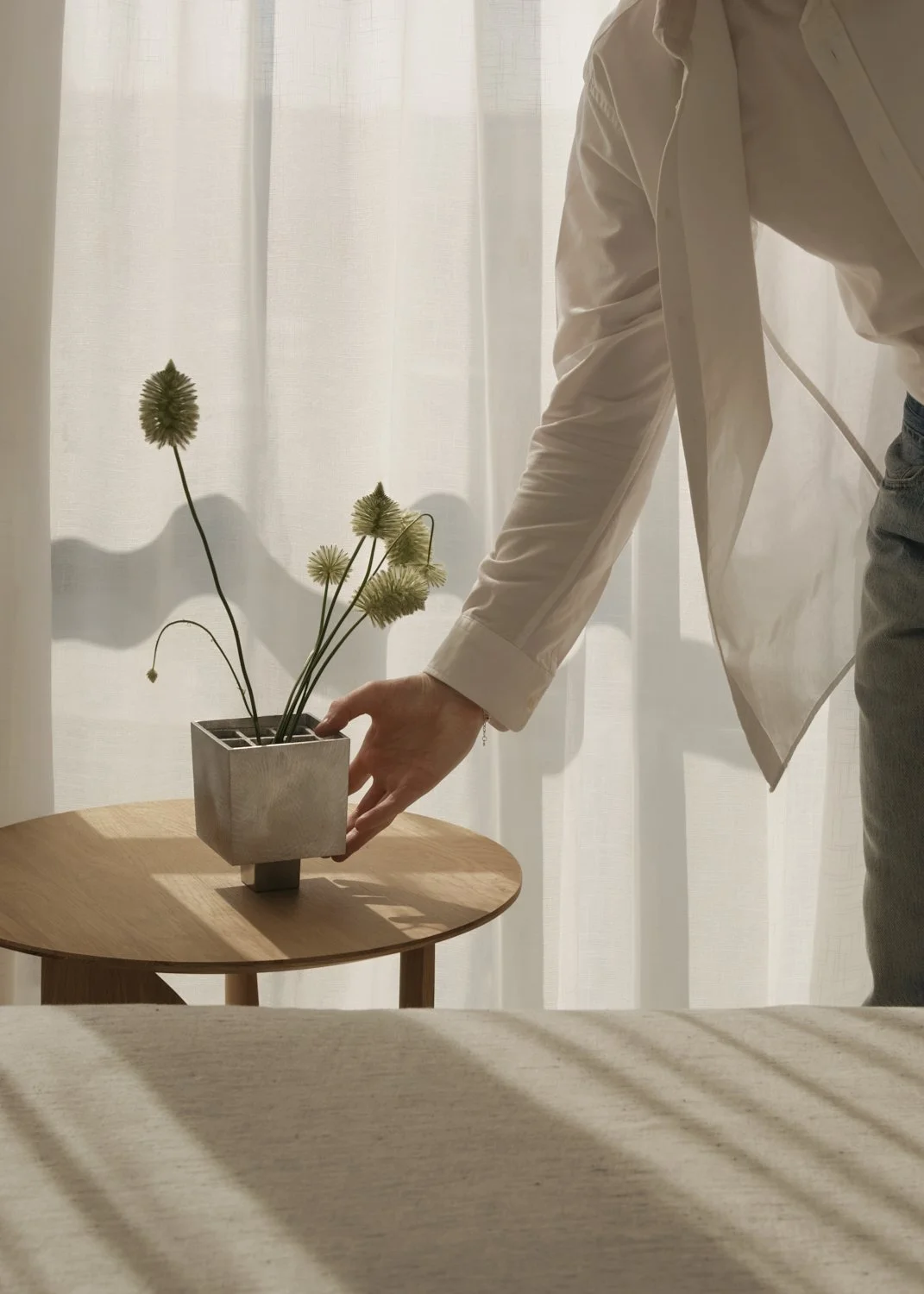 A person in a white shirt arranging a square concrete planter with dried flowers on a round wooden table near a sheer curtain.