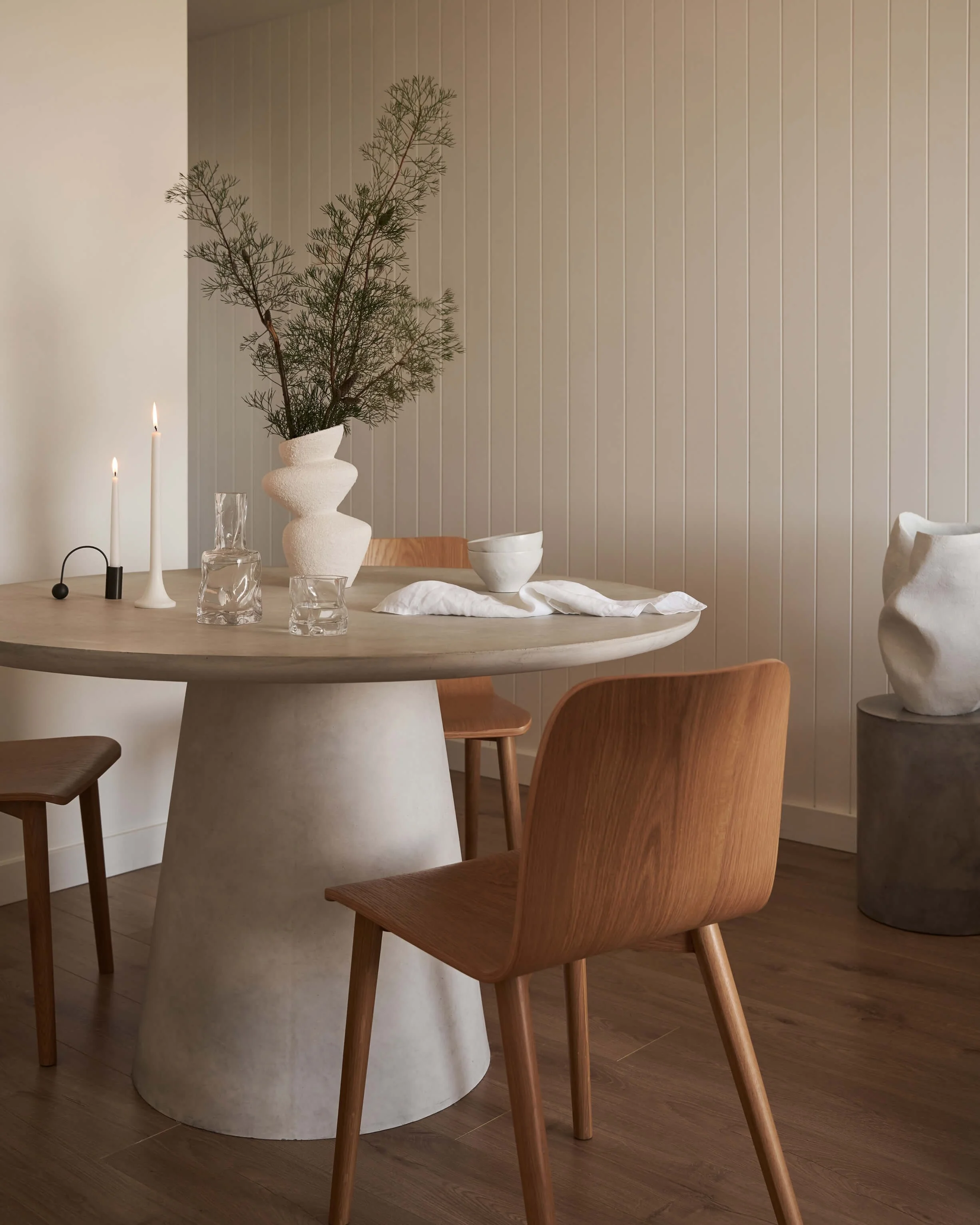 A modern dining room with a round table, wooden chairs, and minimalist decor including a white textured vase with greenery, candles, glassware, and a large white fabric-covered object on a gray pedestal in the corner.