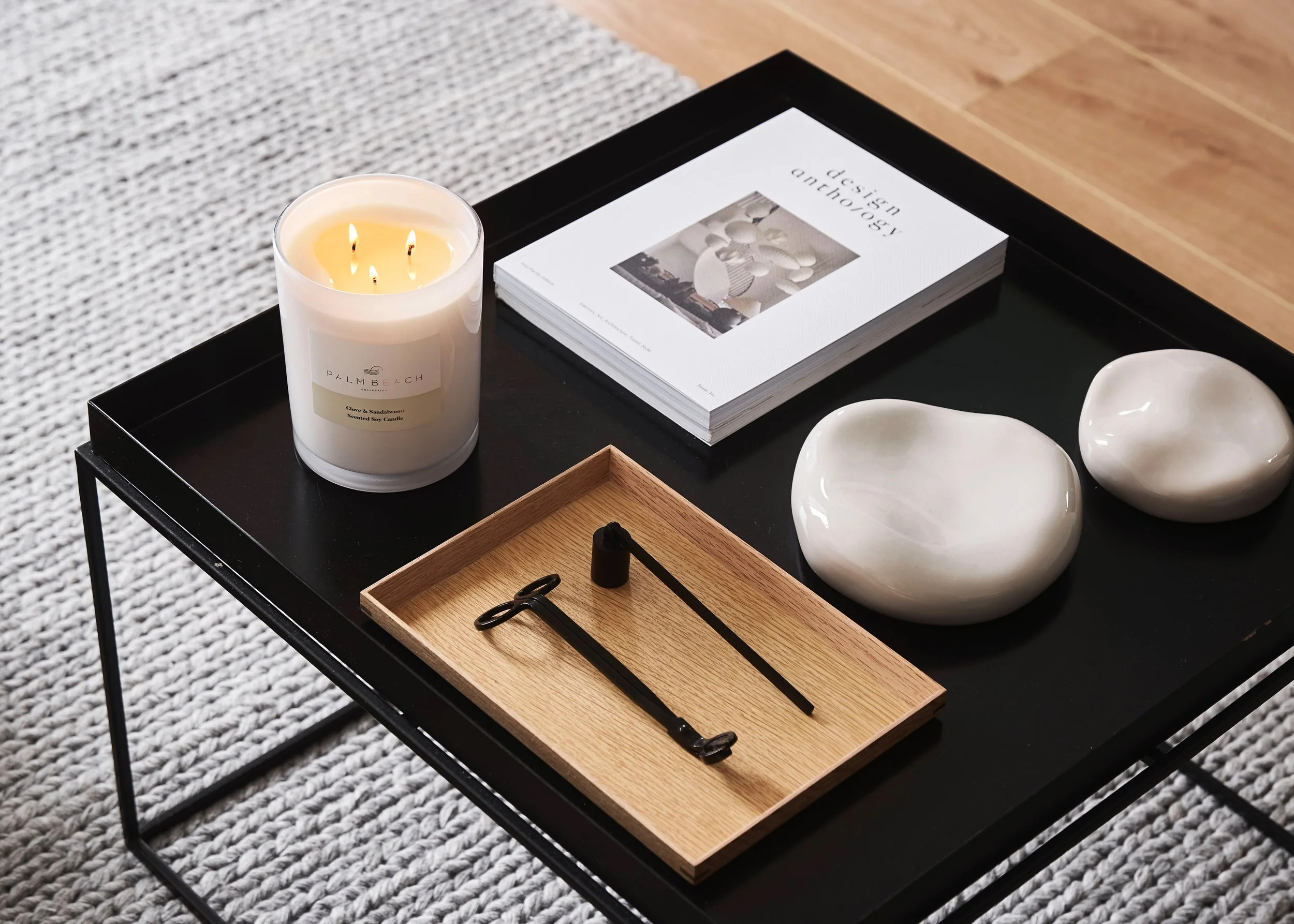 A black side table with a lit white candle, a book titled ‘design anthropology,’ two white ceramic bowls, a wooden tray with a black toothbrush and razor, placed on a grey textured rug on a wooden floor.