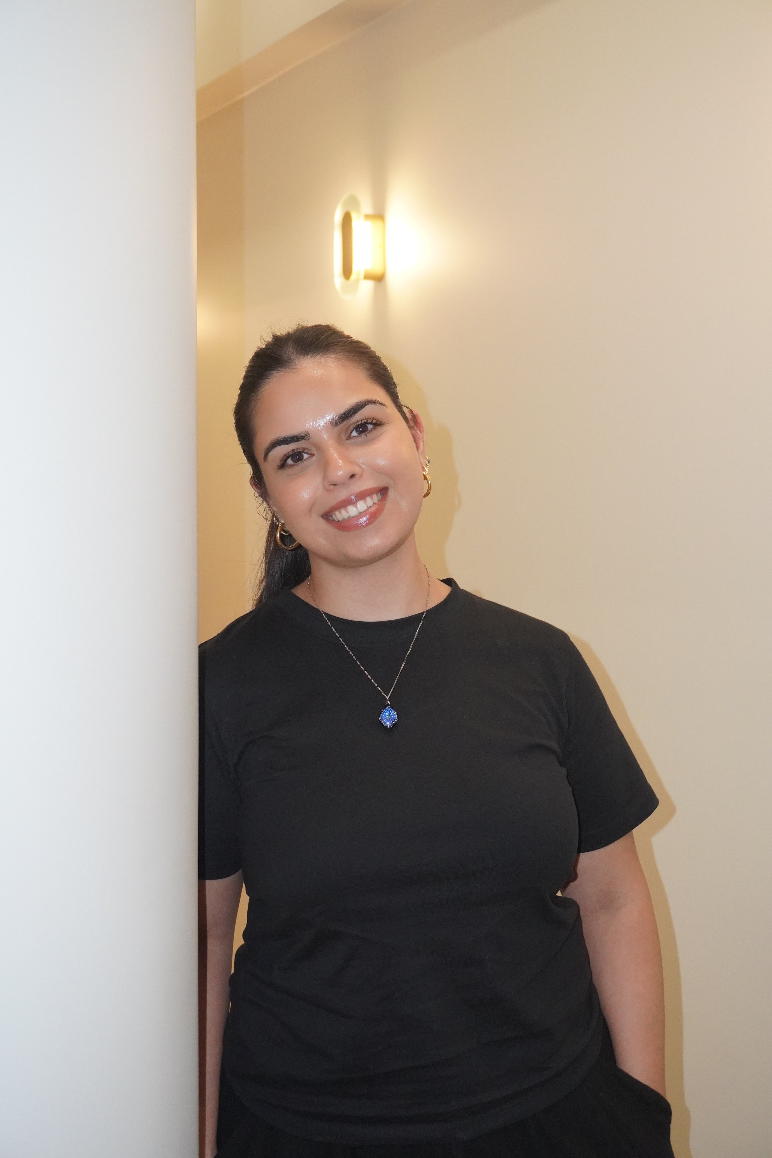 A young woman with dark hair pulled back, wearing a black t-shirt and jewelry, smiling while leaning against a wall with a light fixture.