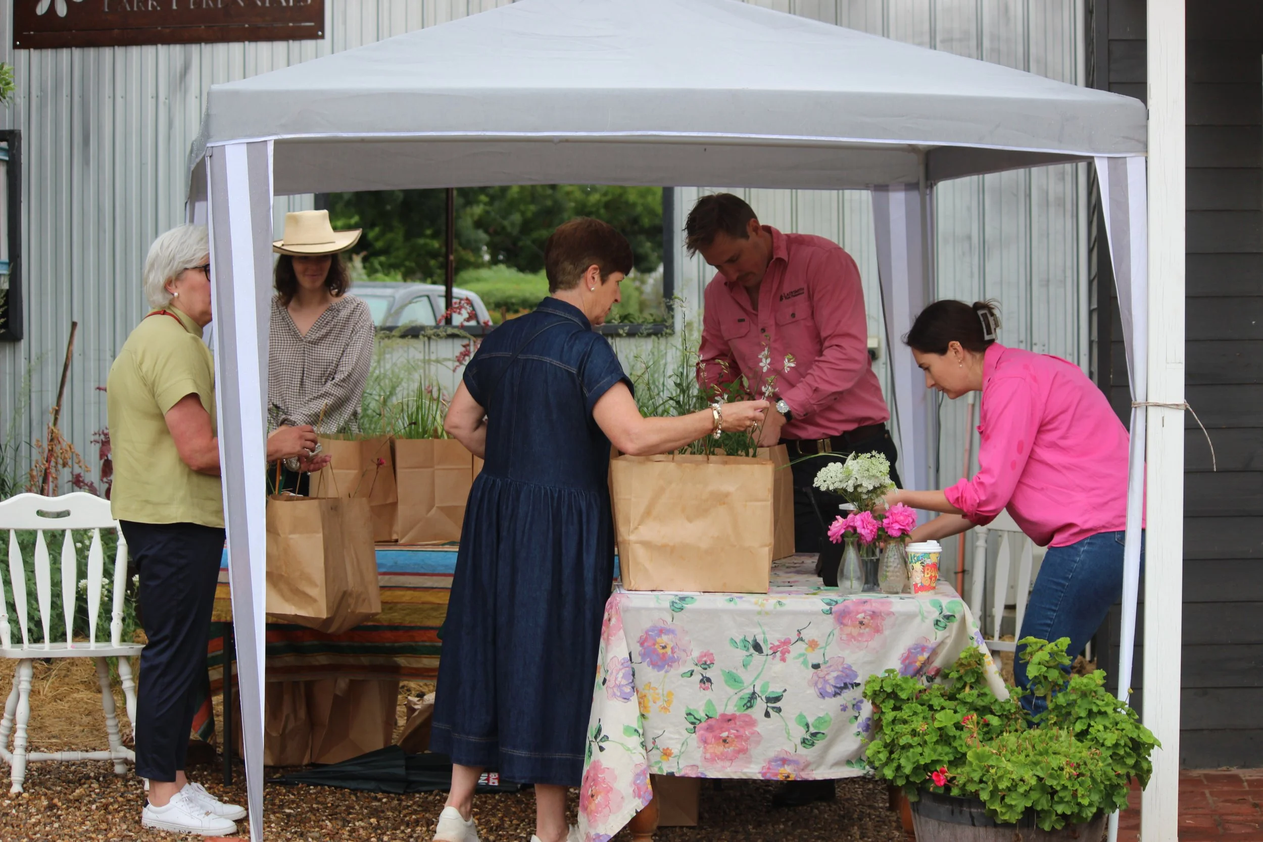 People shopping at a farmers market under a white canopy, with a table of flowers and paper bags.