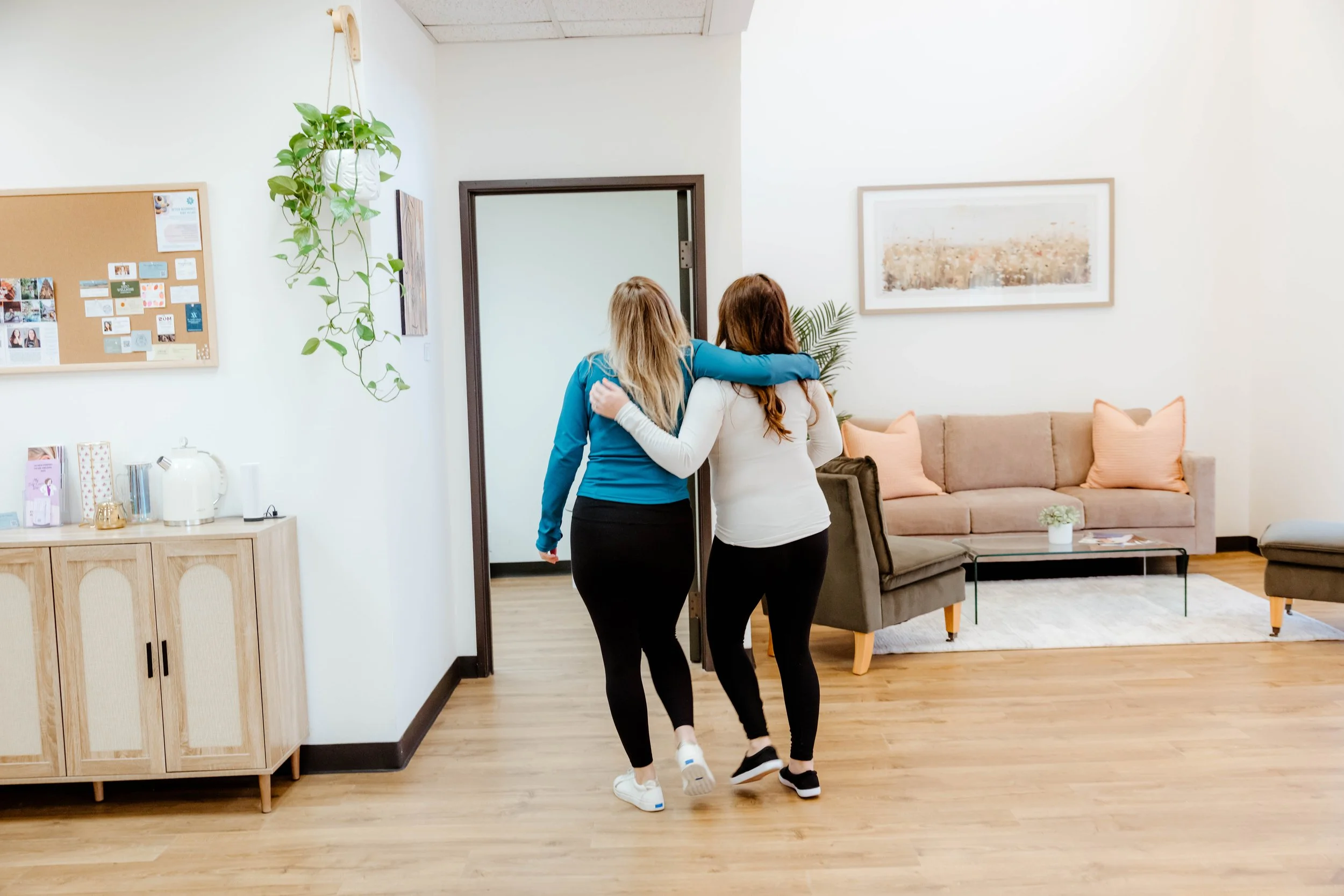 Two women walking arm-in-arm in a cozy living room with a beige couch, wall art, and a wooden sideboard.