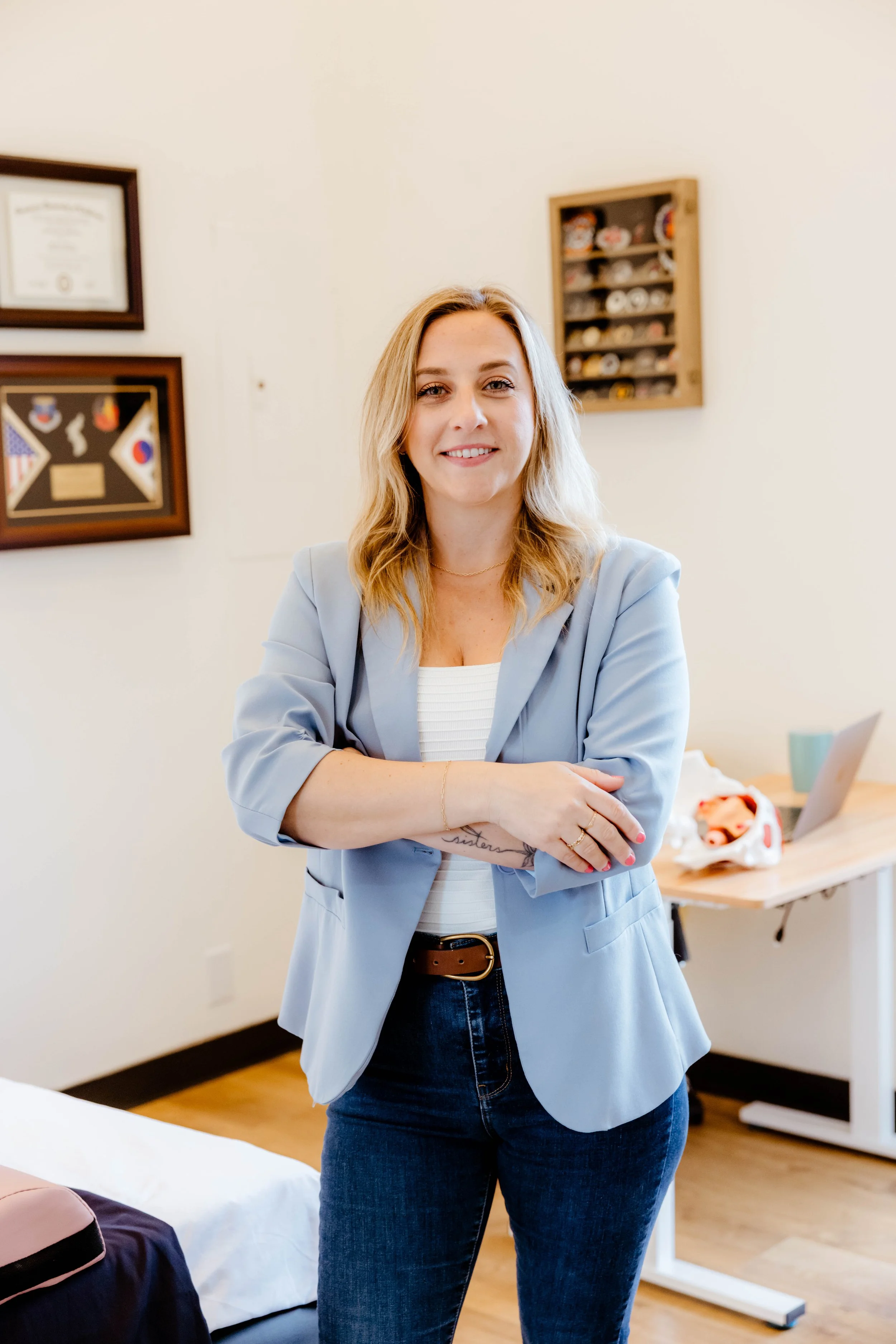 A woman standing with arms crossed in a room with framed art and a small shelf on the wall, dressed in a light blue blazer and jeans, smiling at the camera.