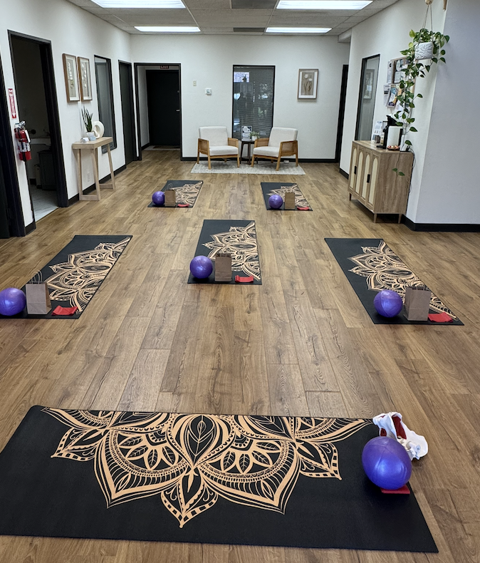 Empty yoga studio with six black and beige mats, each with a purple exercise ball, a small towel, and a bottle of lotion. A seating area with two chairs and a small table is in the background, and a wooden cabinet with a potted plant sits on the right side. The room has light wood flooring and white walls.