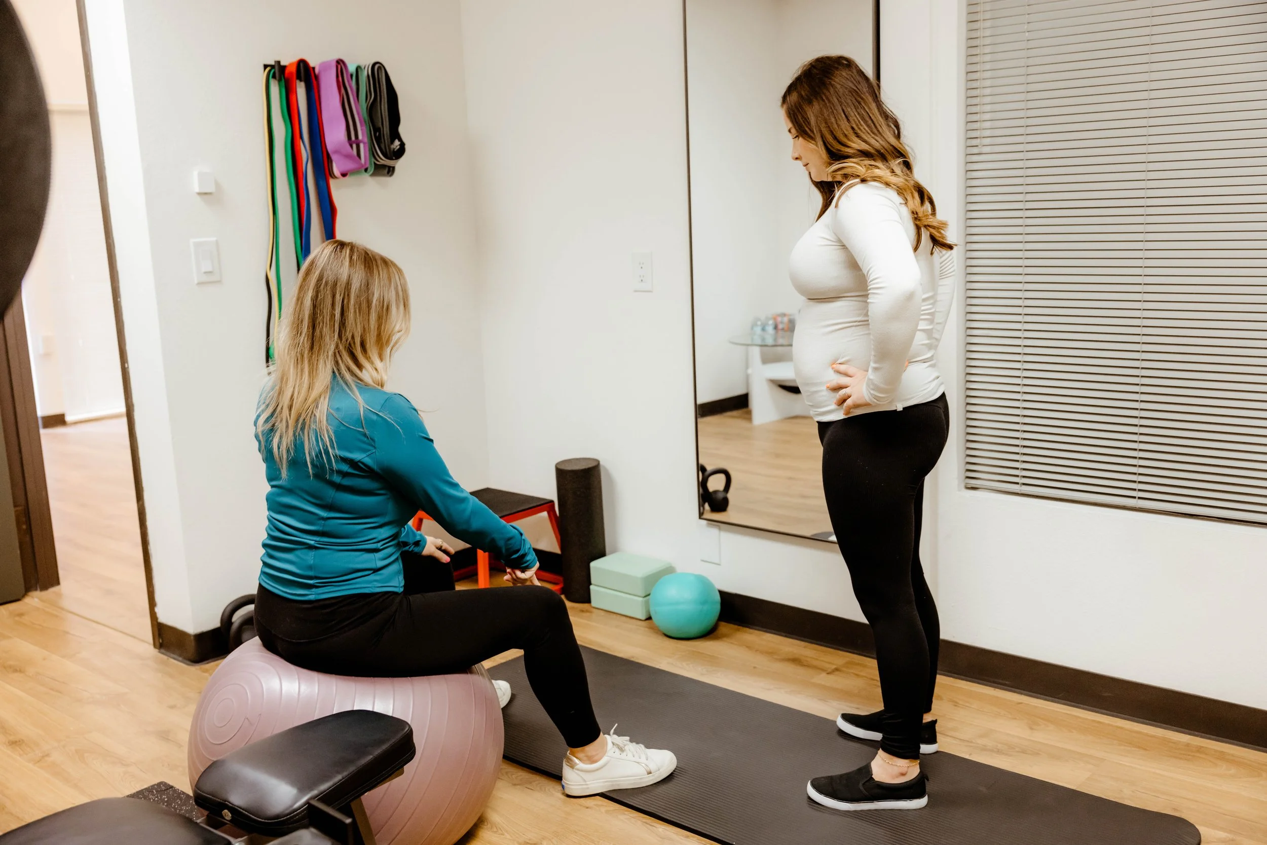 A woman with a pregnant belly stands with hands on hips, looking at a woman sitting on a pink exercise ball, both in a fitness or therapy room with gym equipment and a large mirror.