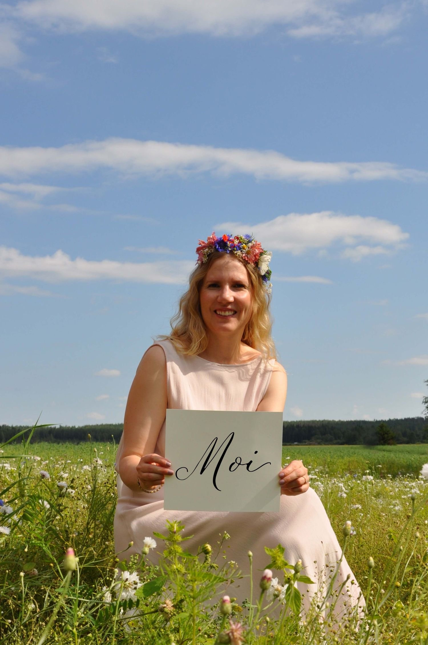 Nainen istuu kukkaniityllä kädessään kyltti, jossa lukee Moi.