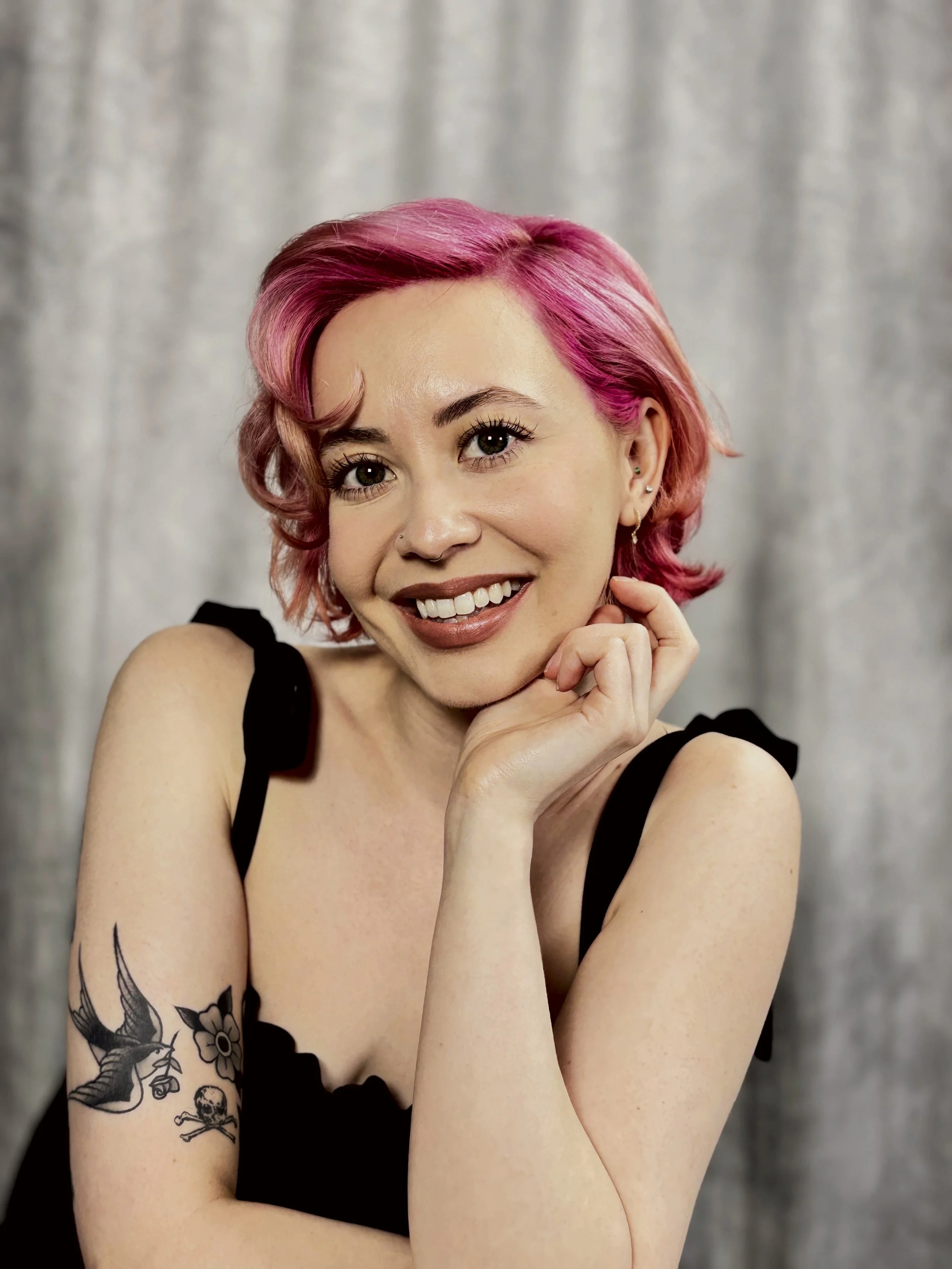 A woman with pink hair and tattoos, smiling, wearing a black sleeveless top, against a neutral background.