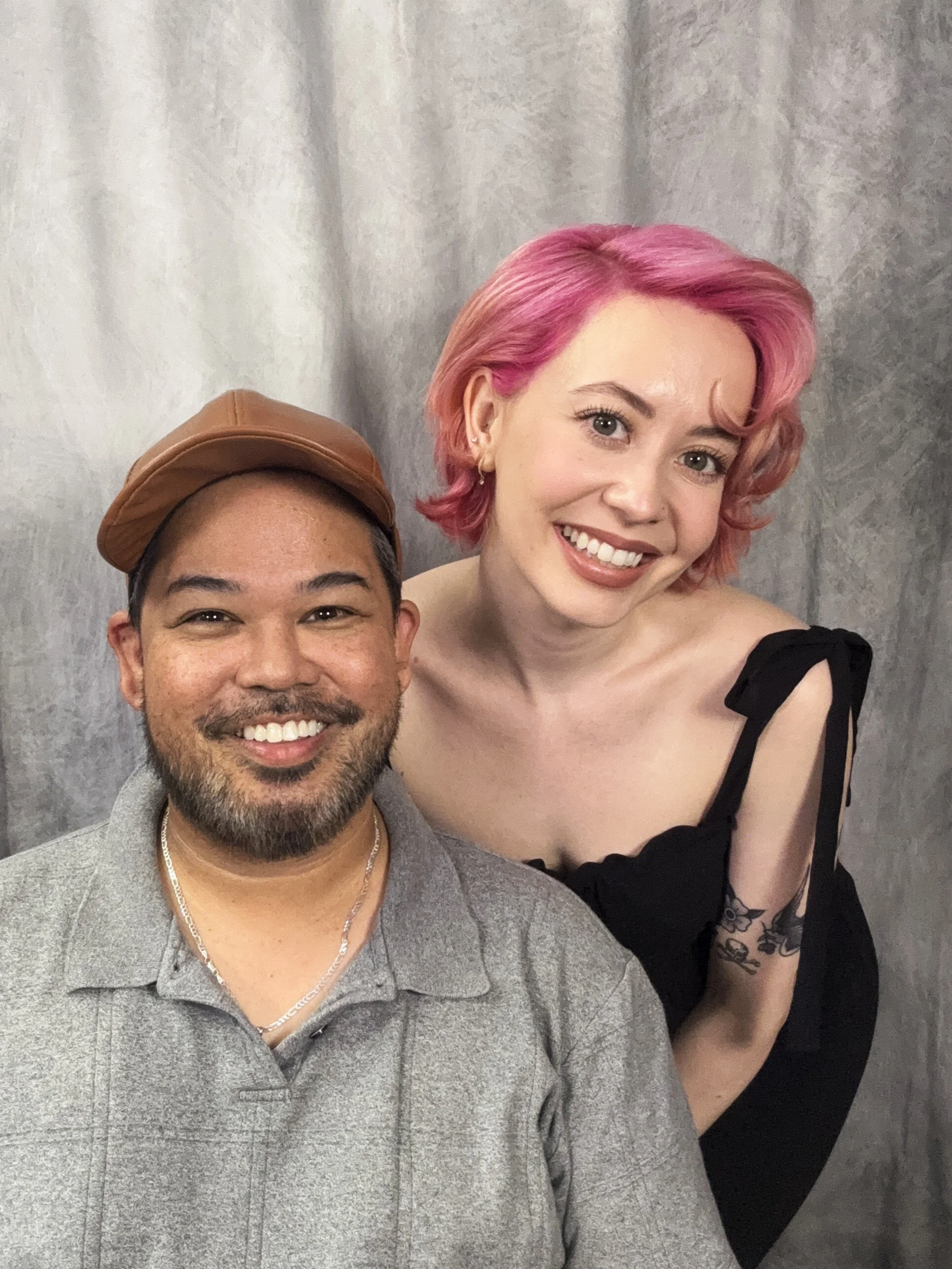A smiling man with a beard, wearing a gray polo shirt, a chain necklace, and a tan cap, standing next to a woman with pink hair, wearing a black dress with ruffled sleeves, and tattoos on her arm, against a gray curtain background.