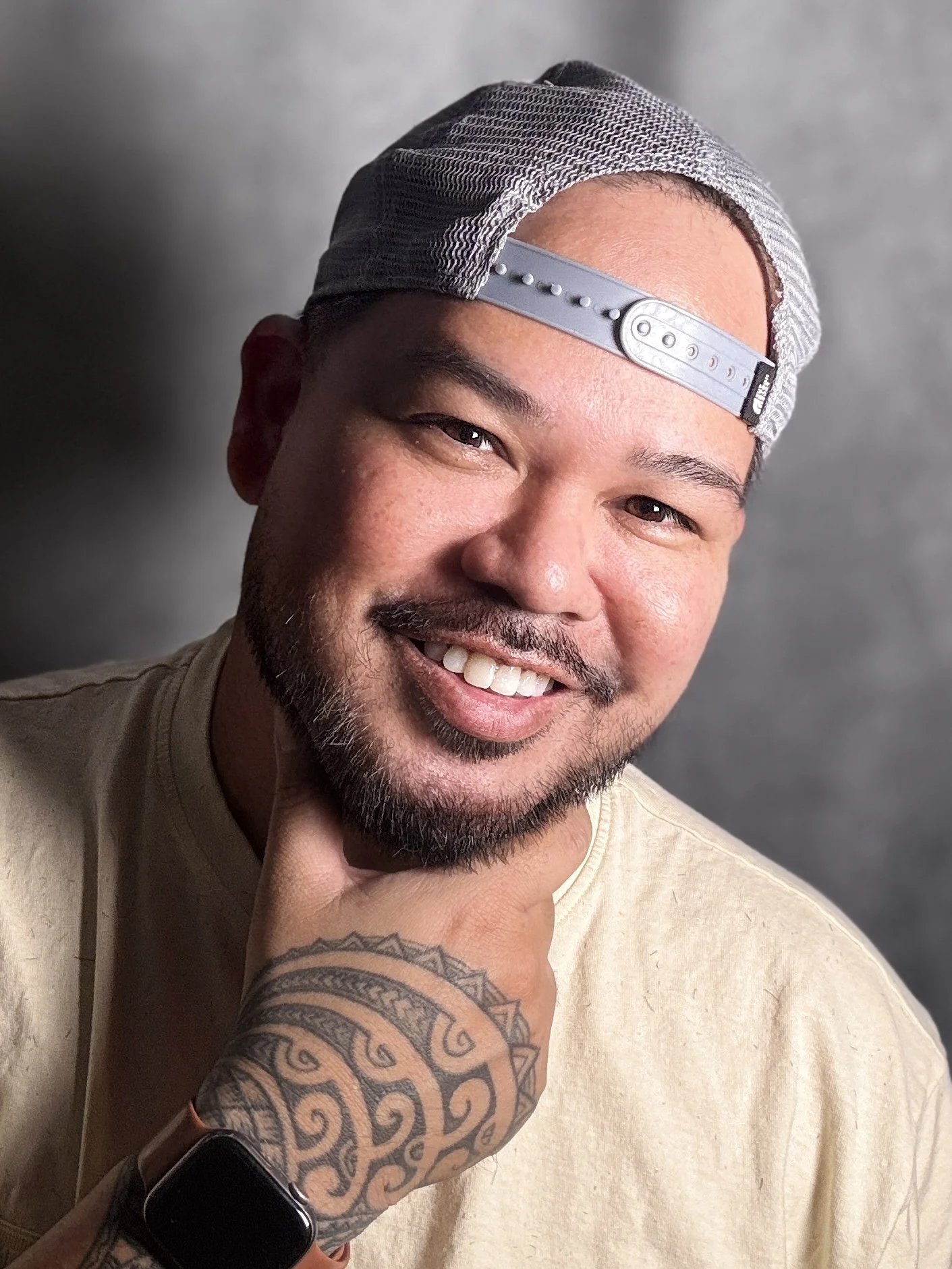 A smiling man with a beard and mustache, wearing a backward gray and black cap, a light-colored T-shirt, and a smartwatch, posing with his hand with a Polynesian tattoo under his chin, against a gray background.