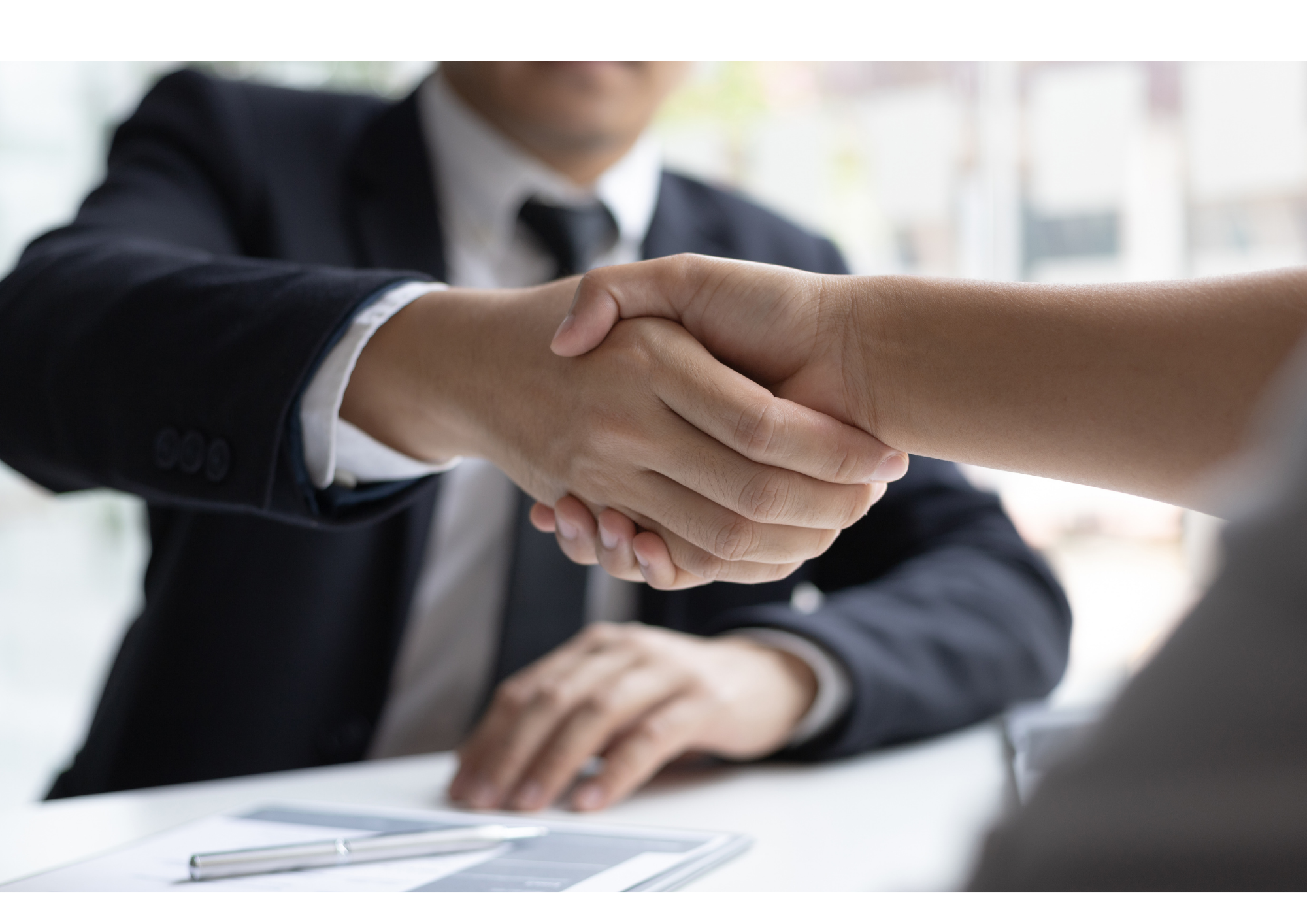 Zwei Personen schütteln sich die Hand bei einem geschäftlichen Treffen, ein Mann in einem schwarzen Anzug und weißem Hemd, im Hintergrund ein moderner, heller Raum.