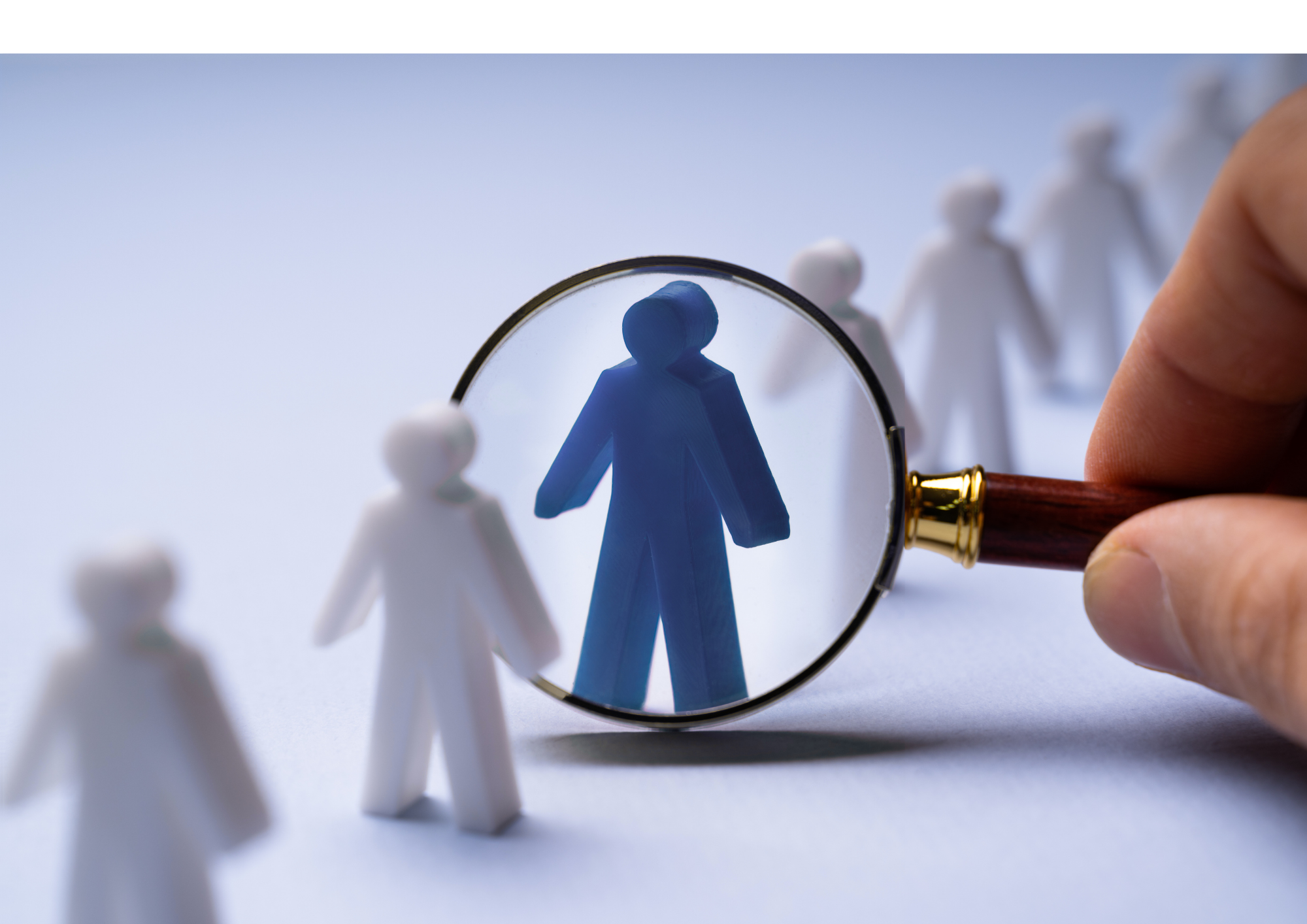 Hand hält eine Lupe, die eine blaue Puppe unter einer Reihe von weißen Puppen vergrößert zeigt, symbolisiert die Suche oder Auswahl einer Person.